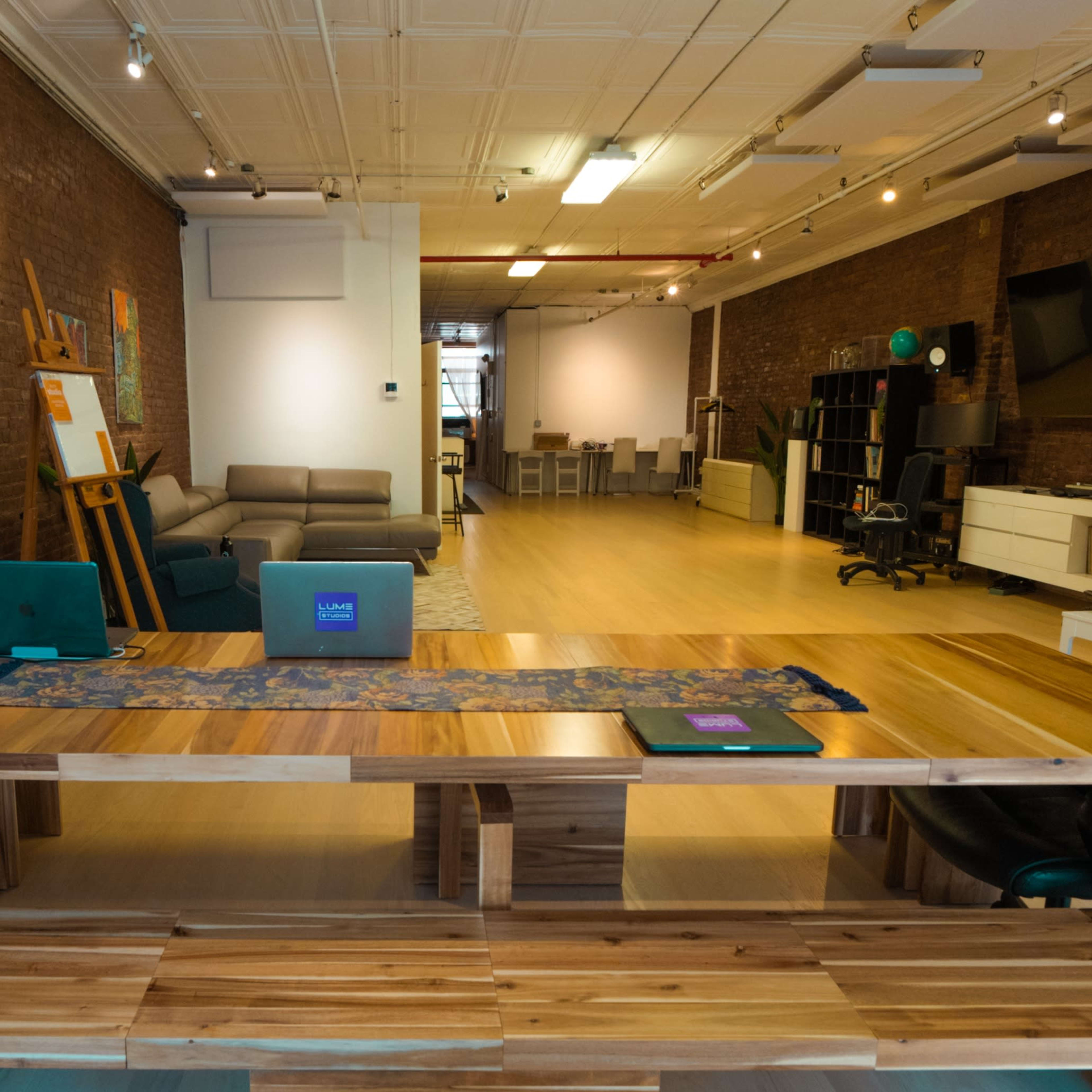 A spacious, modern studio features a large wooden table, a sleek laptop on it, and exposed brick walls, with seating and various workspaces in the background.