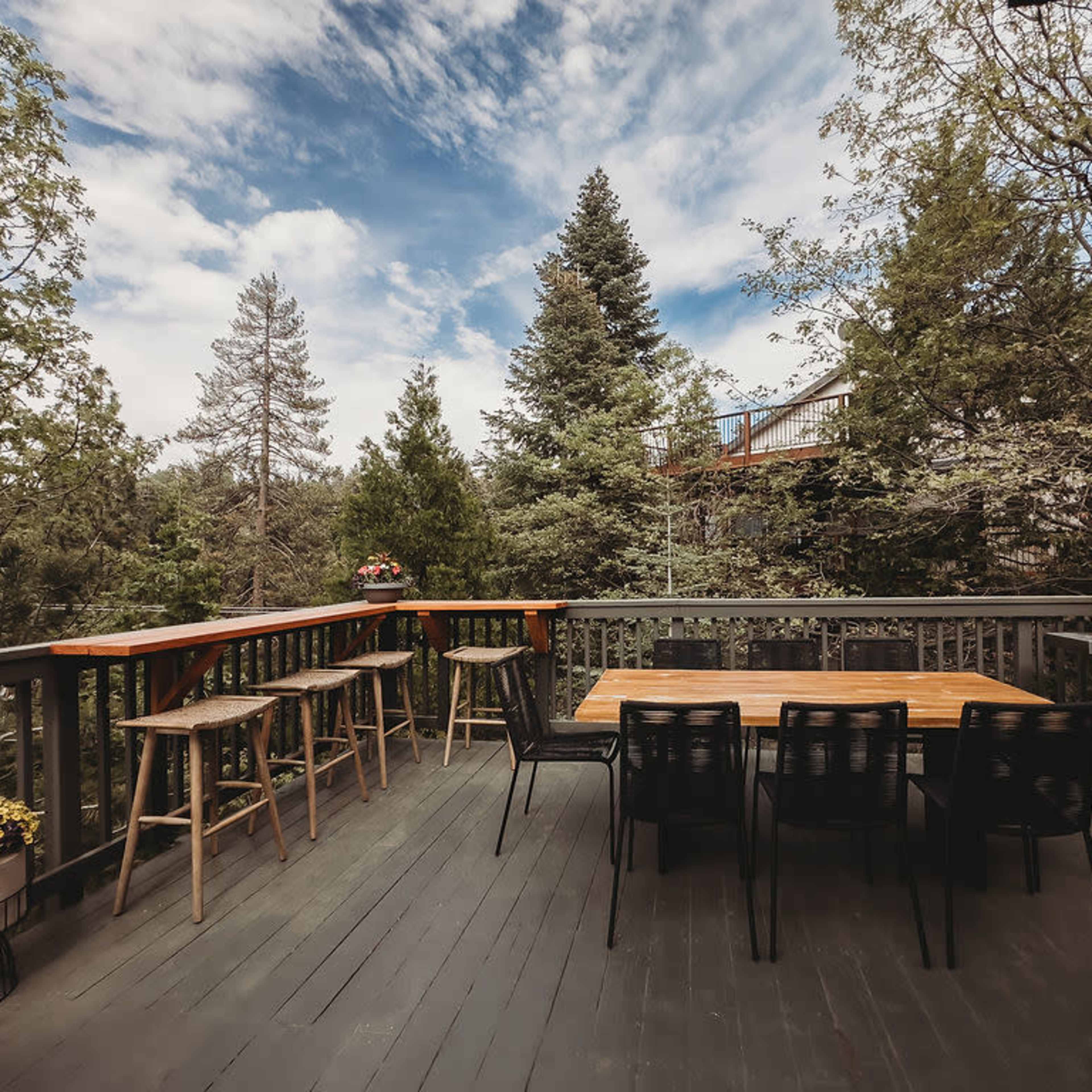 A wooden deck features a dining table with chairs, bar stools, and a grill, surrounded by trees and a cloudy sky.