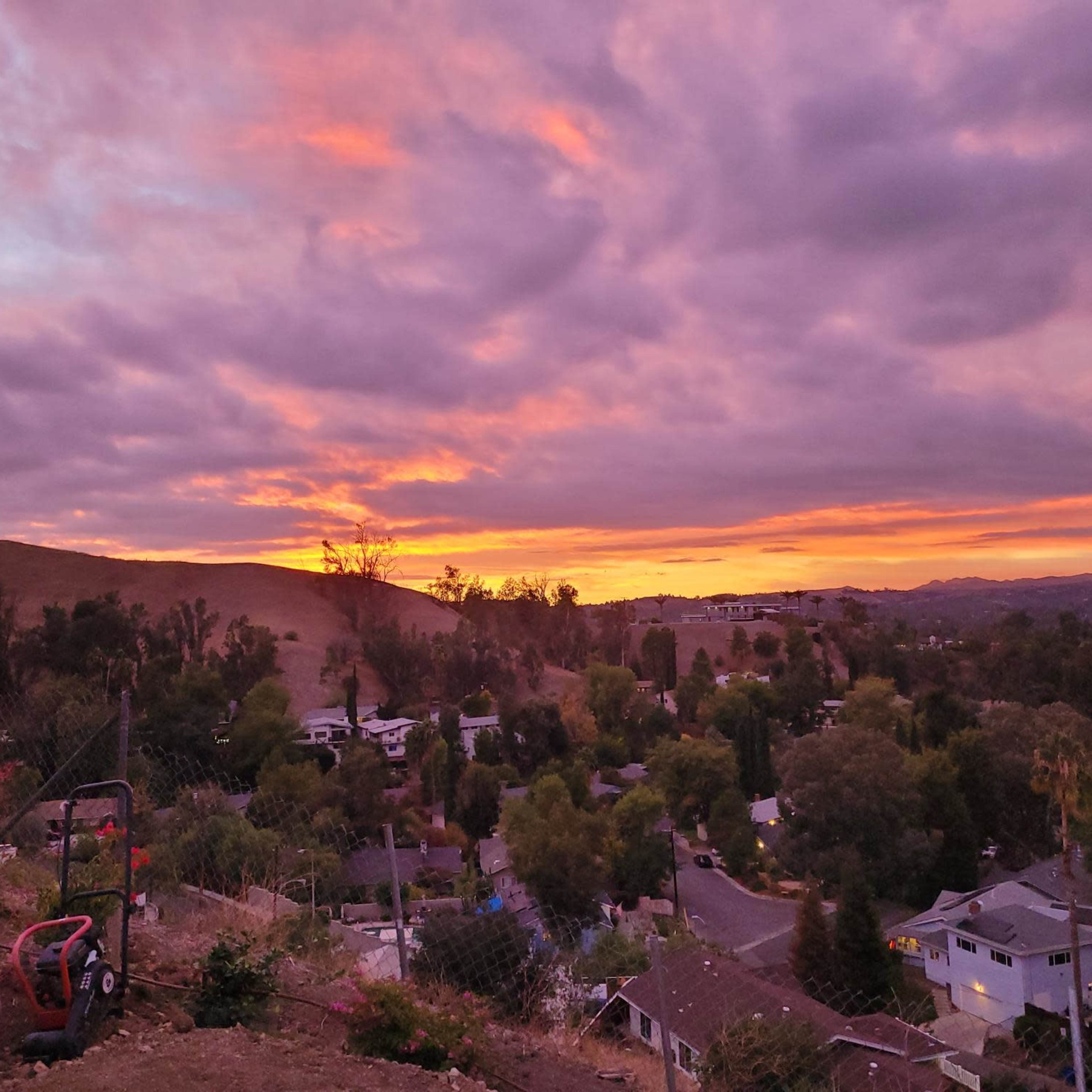 The scene shows a sunset over a residential neighborhood, with hills in the background and a sky filled with purple and orange hues.