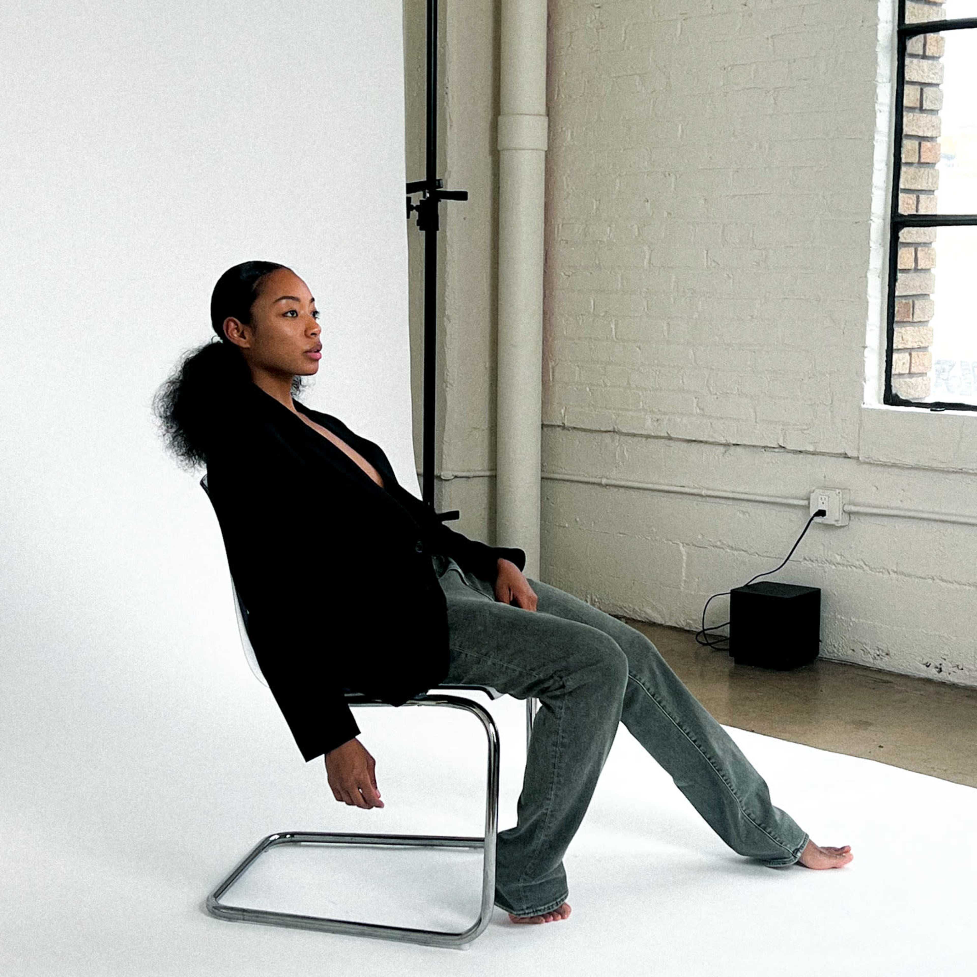 A person sits on a metal chair in a minimalist studio space, wearing a black blazer and gray pants, with one foot bare.