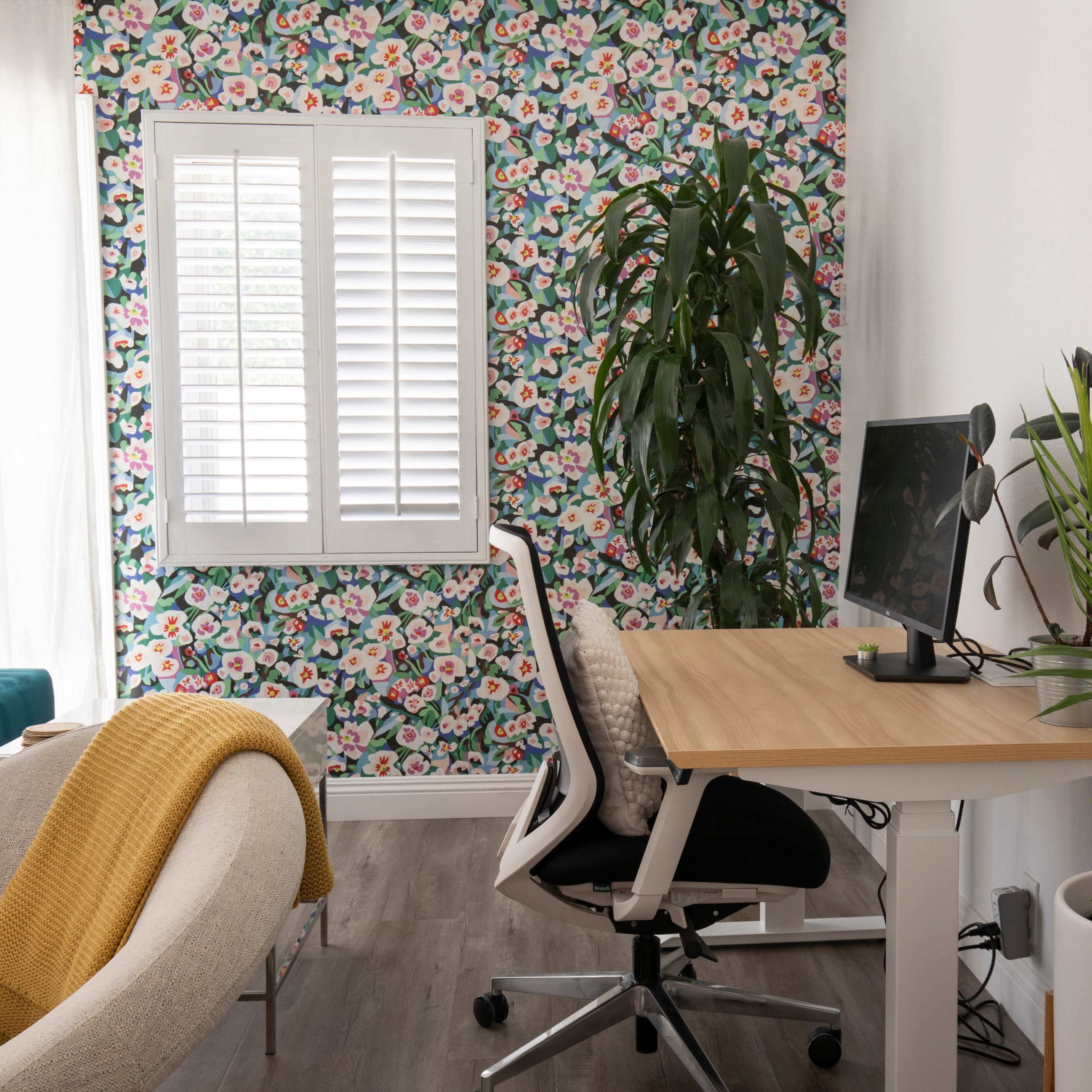 A home office area features a desk with a computer, an ergonomic chair, and a houseplant beside a window, while a colorful floral wallpaper decorates the wall.