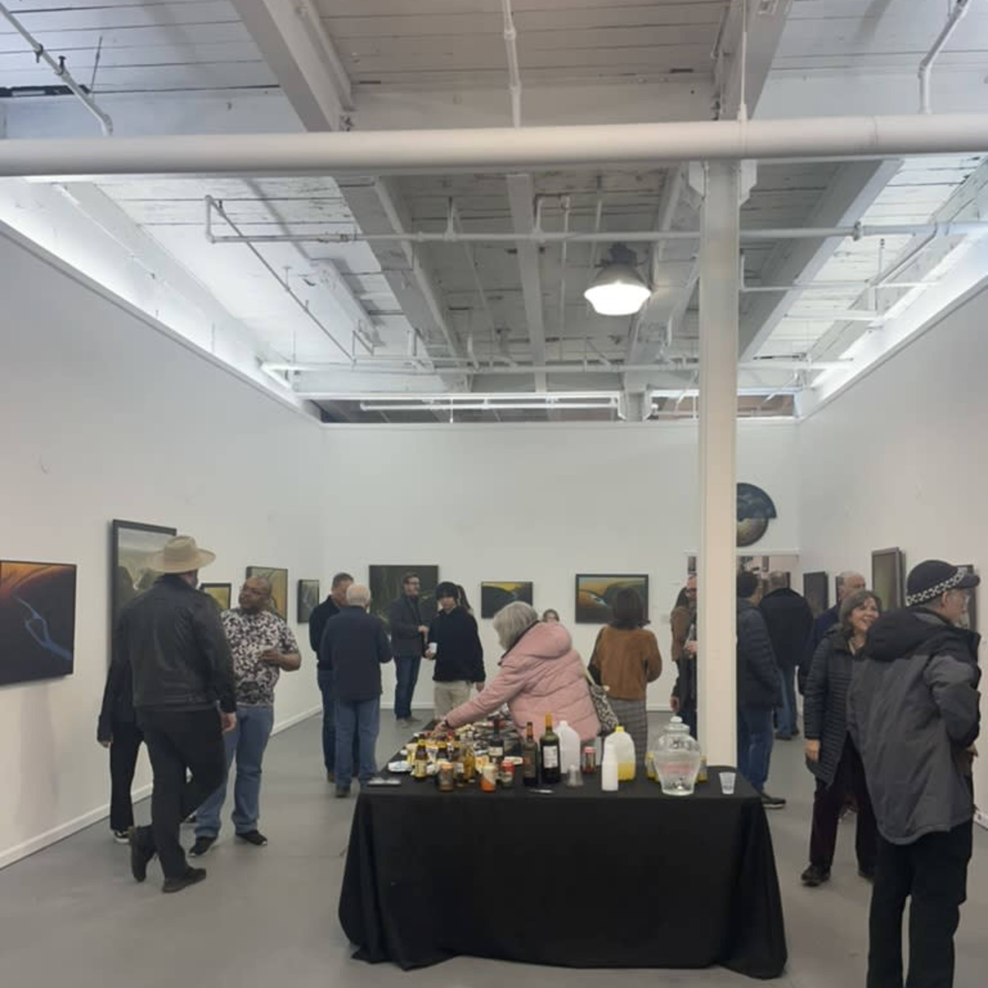 A group of people mingles in an art gallery featuring several paintings on white walls, with a table of refreshments in the foreground.