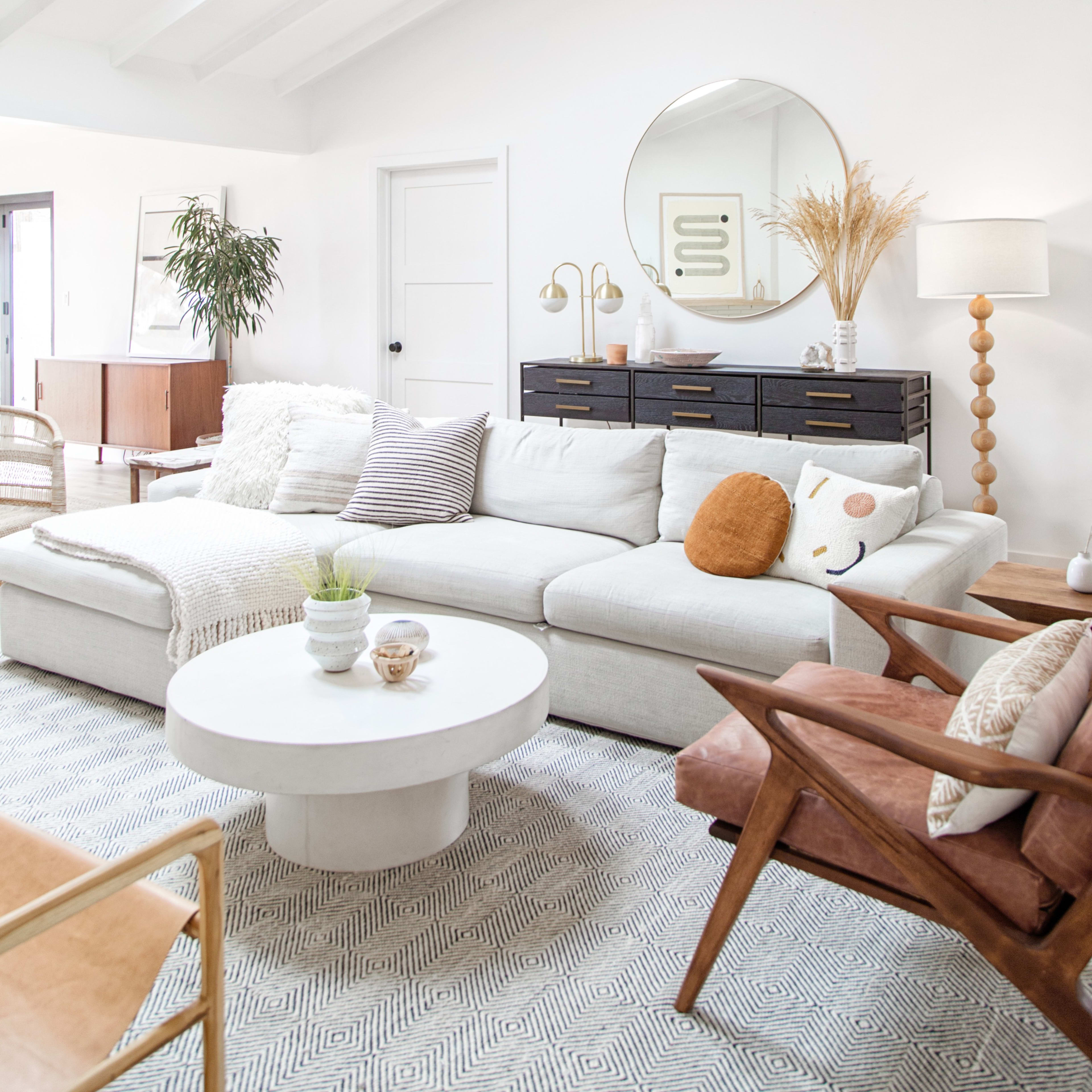 A modern living room with a white sofa, a round coffee table, and a large mirror, featuring natural light from a skylight and various decorative elements.