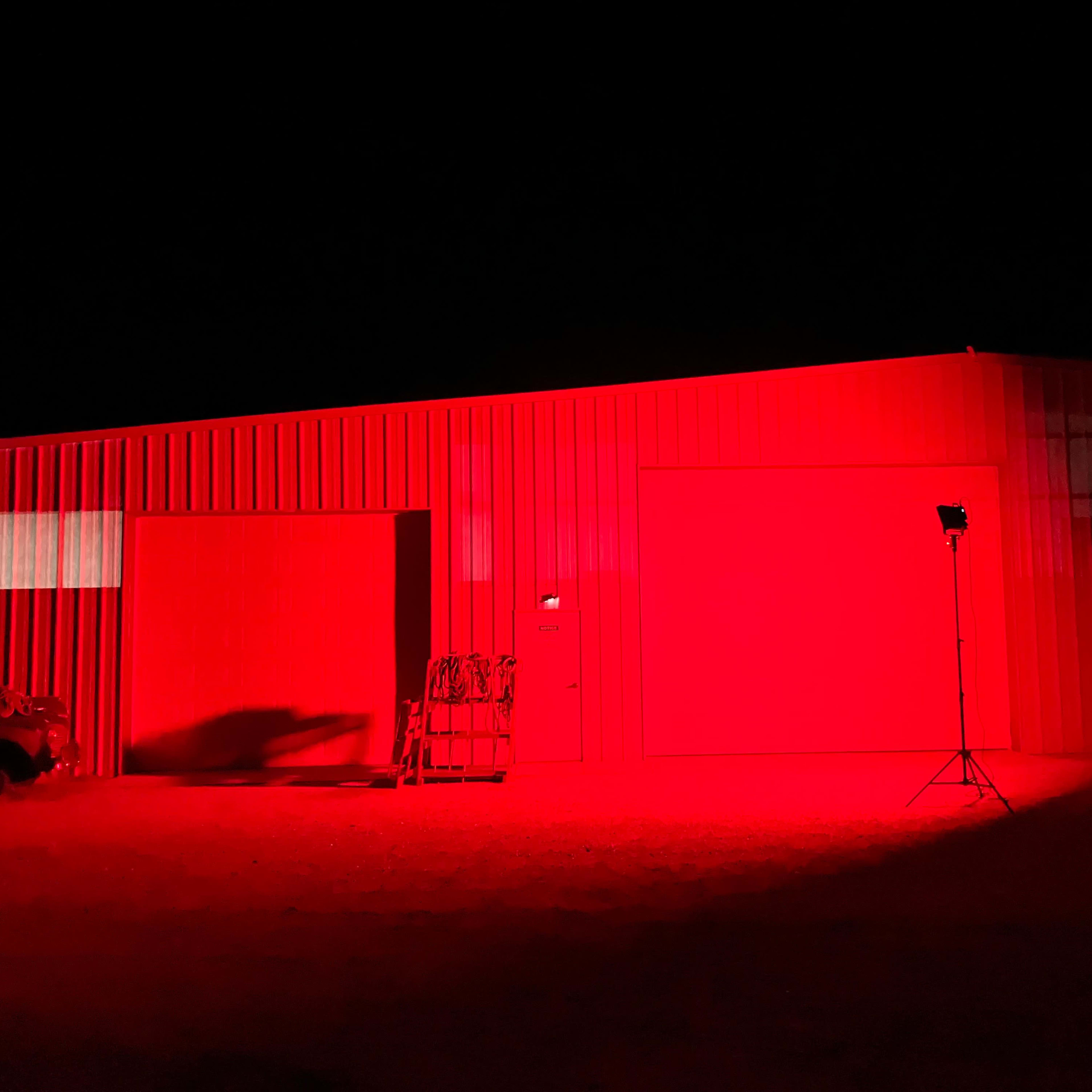 A metal building is illuminated with red light at night, highlighting two large doors and a nearby tripod-mounted light.