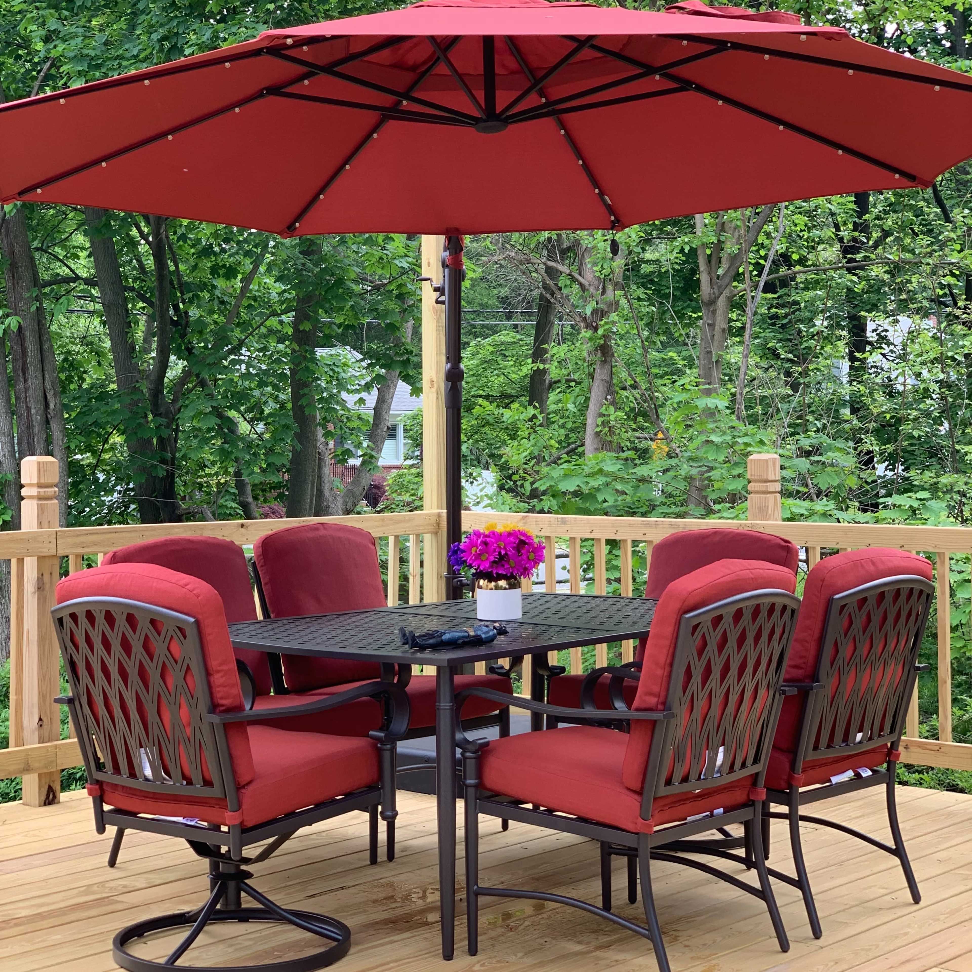 A red patio umbrella shades a set of six cushioned chairs around a black metal table on a wooden deck surrounded by greenery.