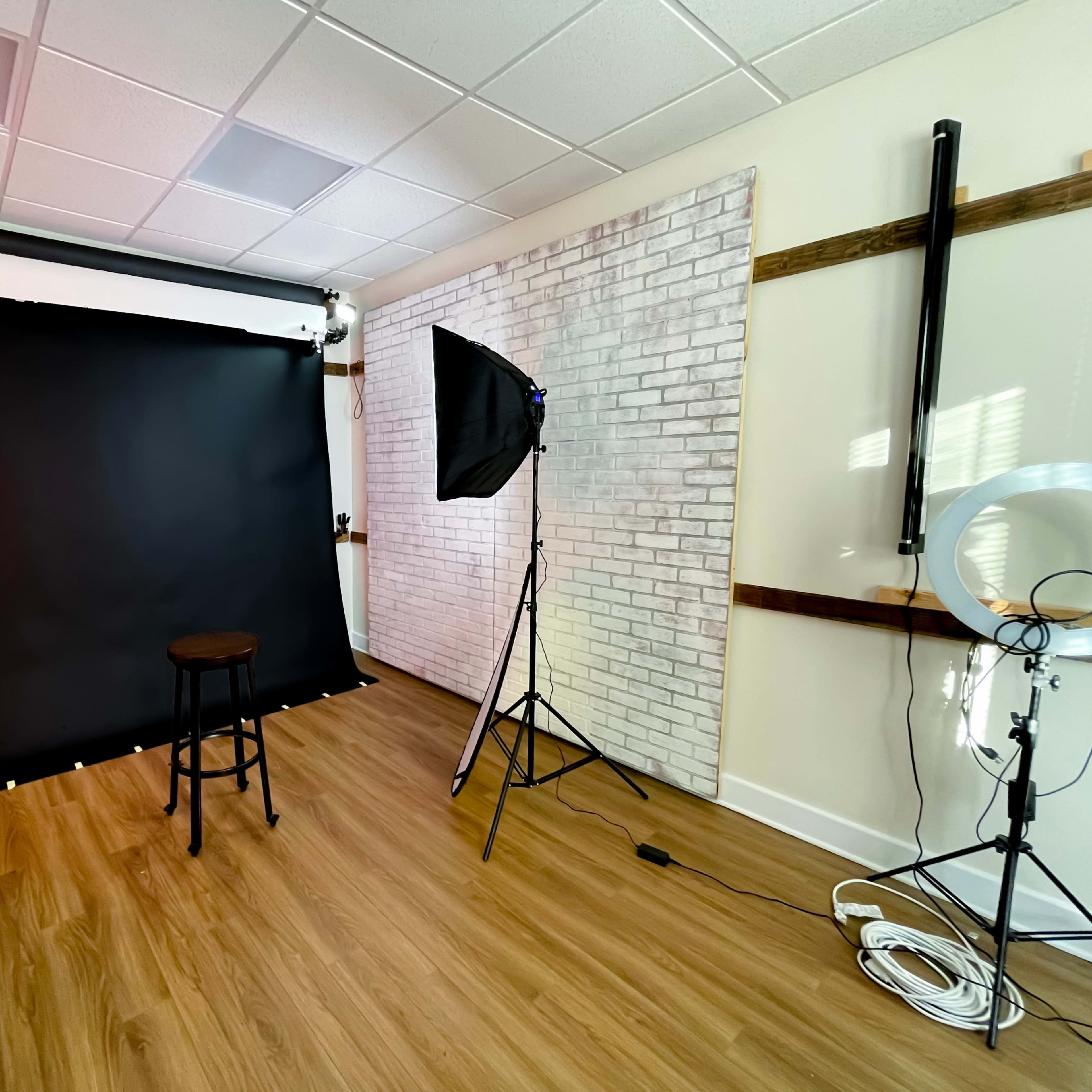 A photography studio with a black backdrop, a stool, two softbox lights, and a circular light setup in a room featuring wooden accents and a brick wall.