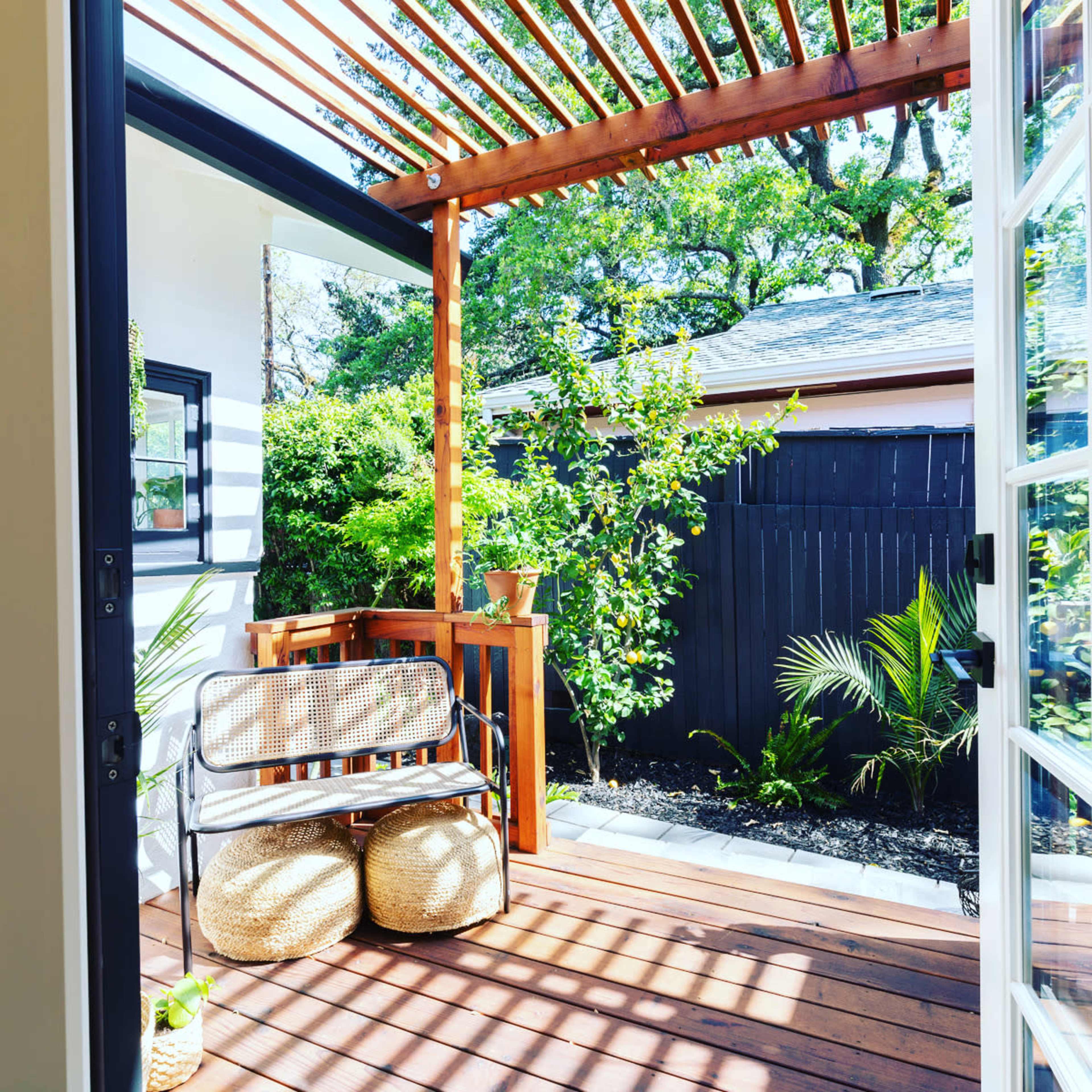 A wooden deck with a bench and two round poufs is framed by open French doors, featuring lush greenery and a shaded area from a slatted pergola overhead.