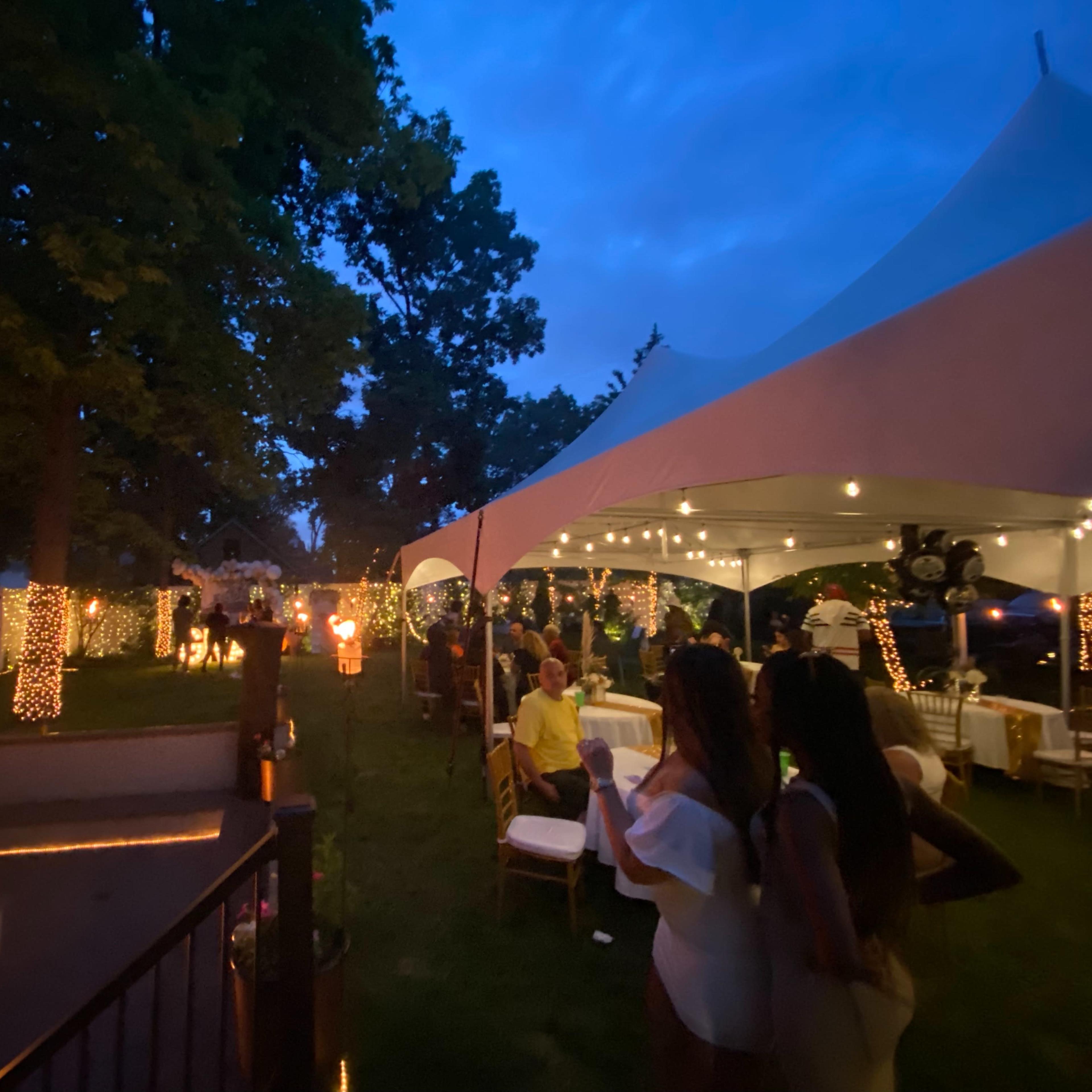 A festive outdoor gathering features a lit gazebo with string lights, a fire pit, and guests socializing under a large tent as dusk sets in.