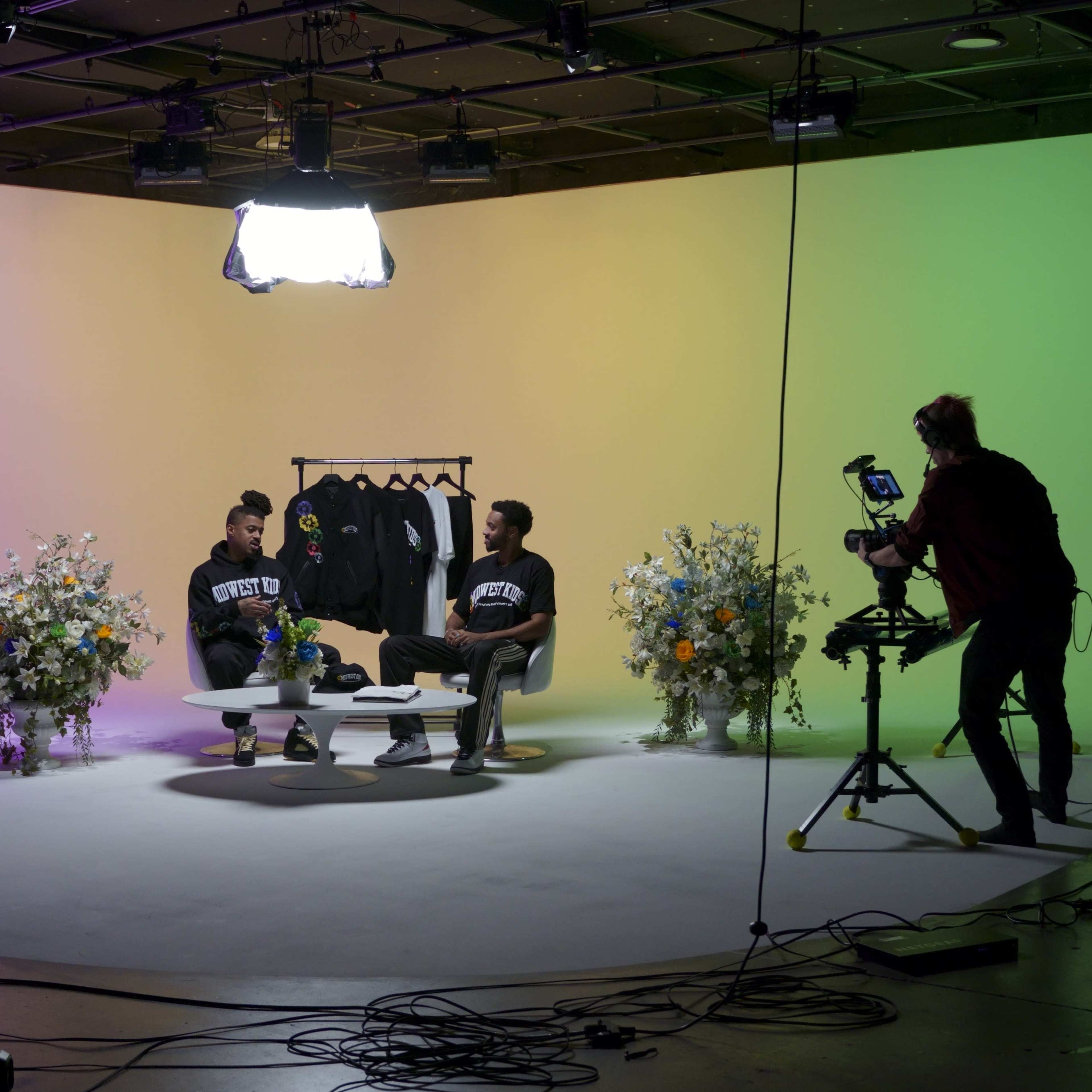 Two individuals sit on a set with colorful backgrounds and floral arrangements, while cameras and lighting equipment surround them.