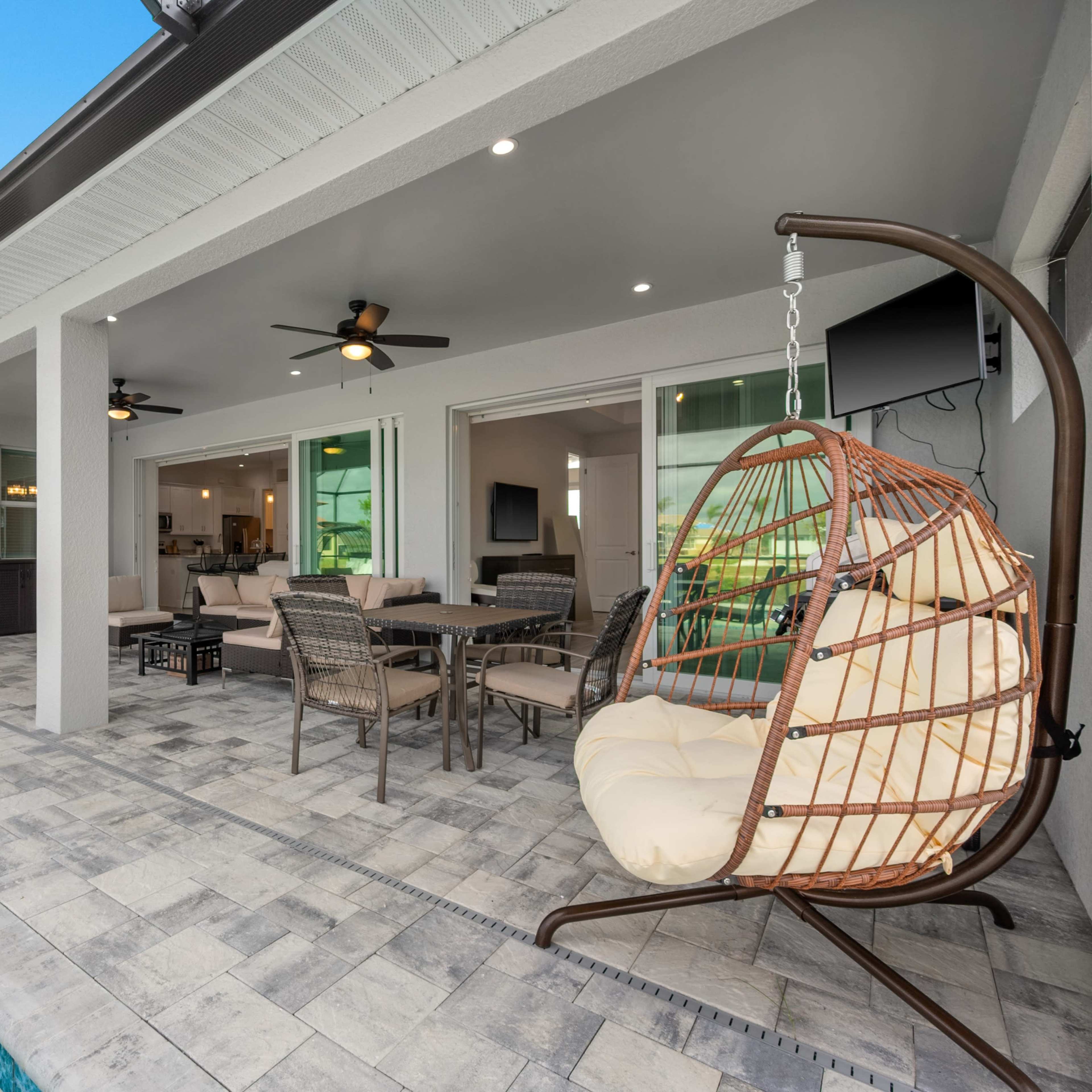 A modern outdoor patio area featuring a swinging chair, a dining table, and a swimming pool, all under a structured roof.