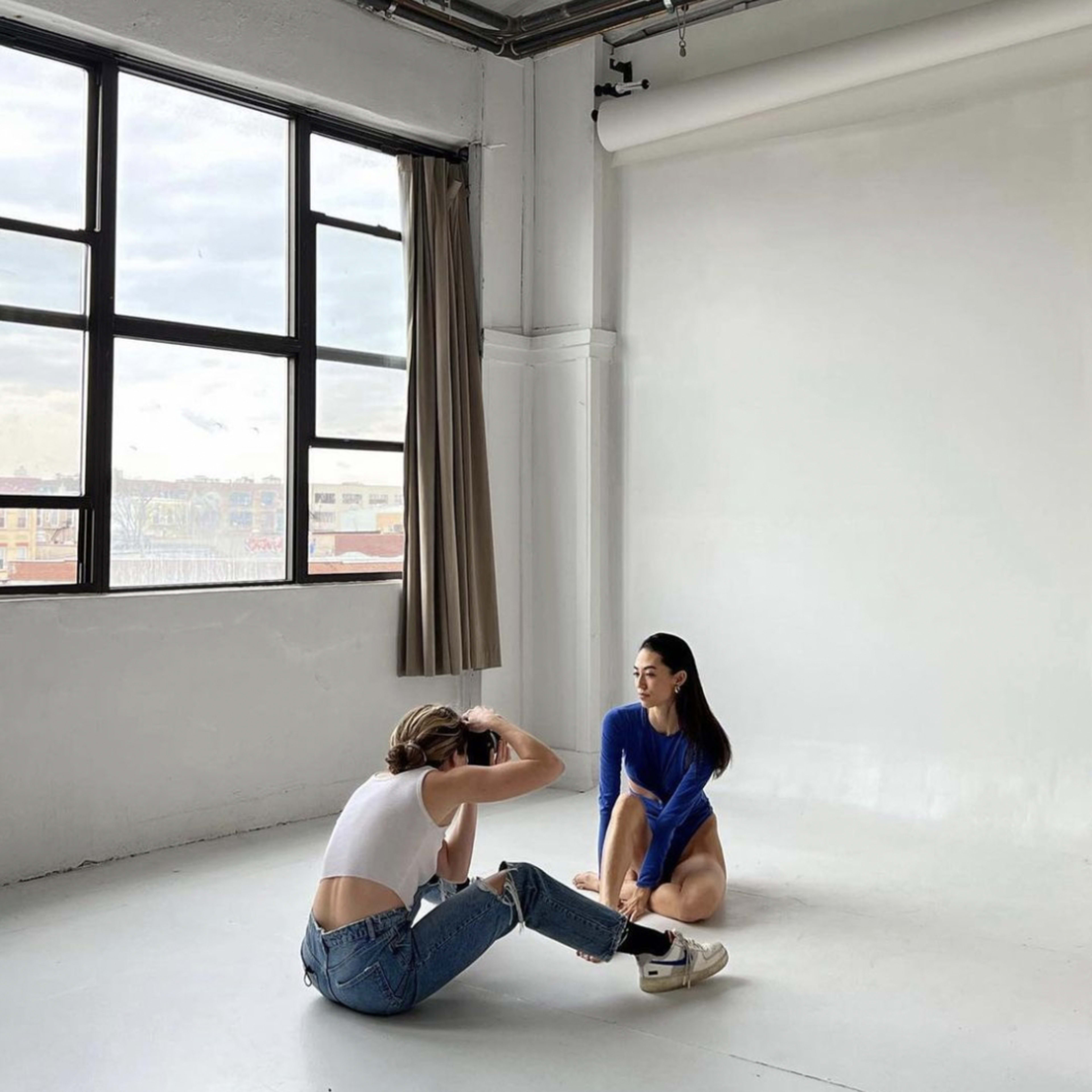 A photographer crouches on the floor in a studio, capturing images of a model sitting against a plain wall.