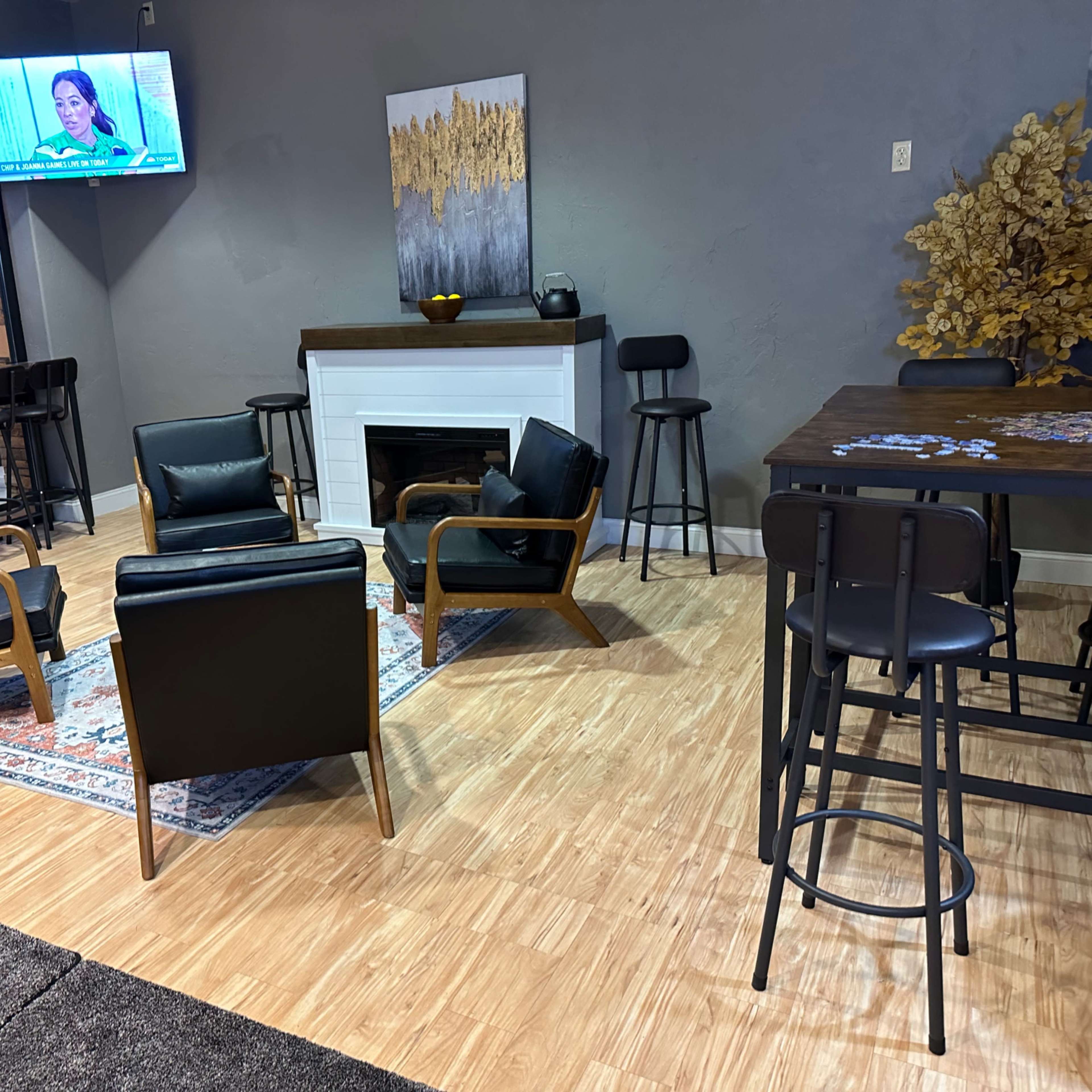 The scene features a cozy lounge area with four black chairs arranged around a small rug, a fireplace, and a bar table in the background, while a television displays a program on the wall.