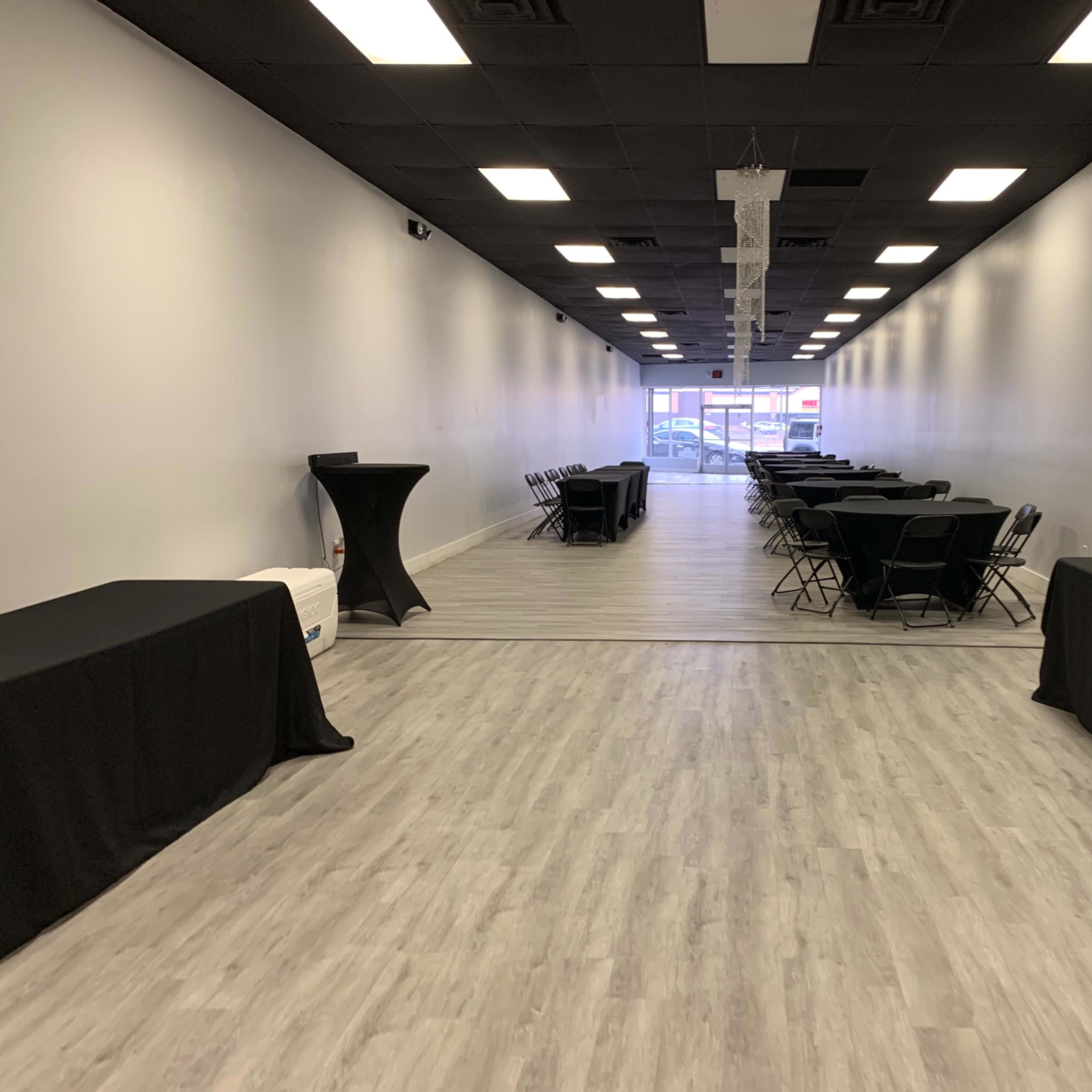 A spacious indoor venue with rows of black tables and chairs set up along a light-colored wooden floor, leading to a large entrance at the far end.