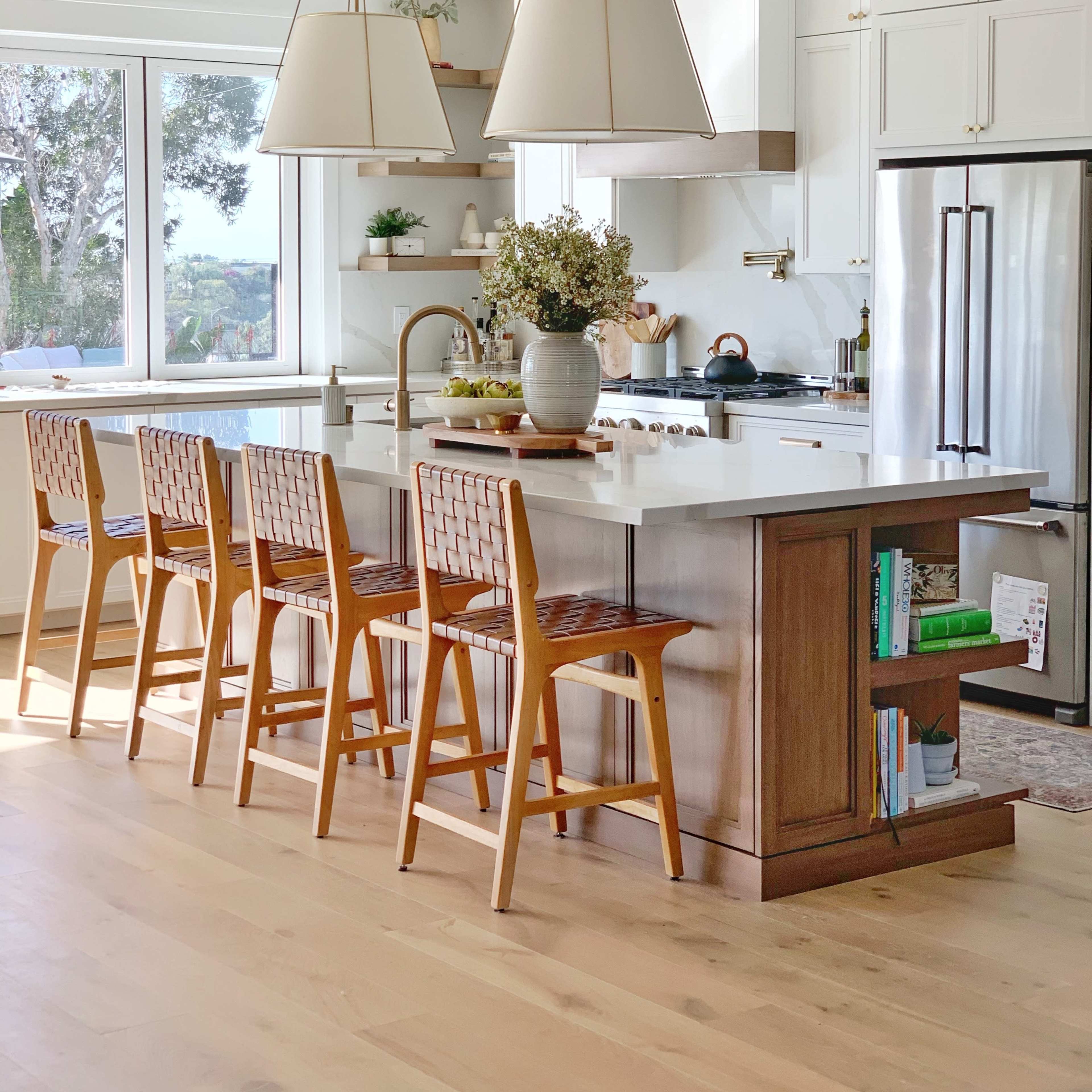A modern kitchen features a central island with bar stools, pendant lights, and large windows allowing natural light to enter.