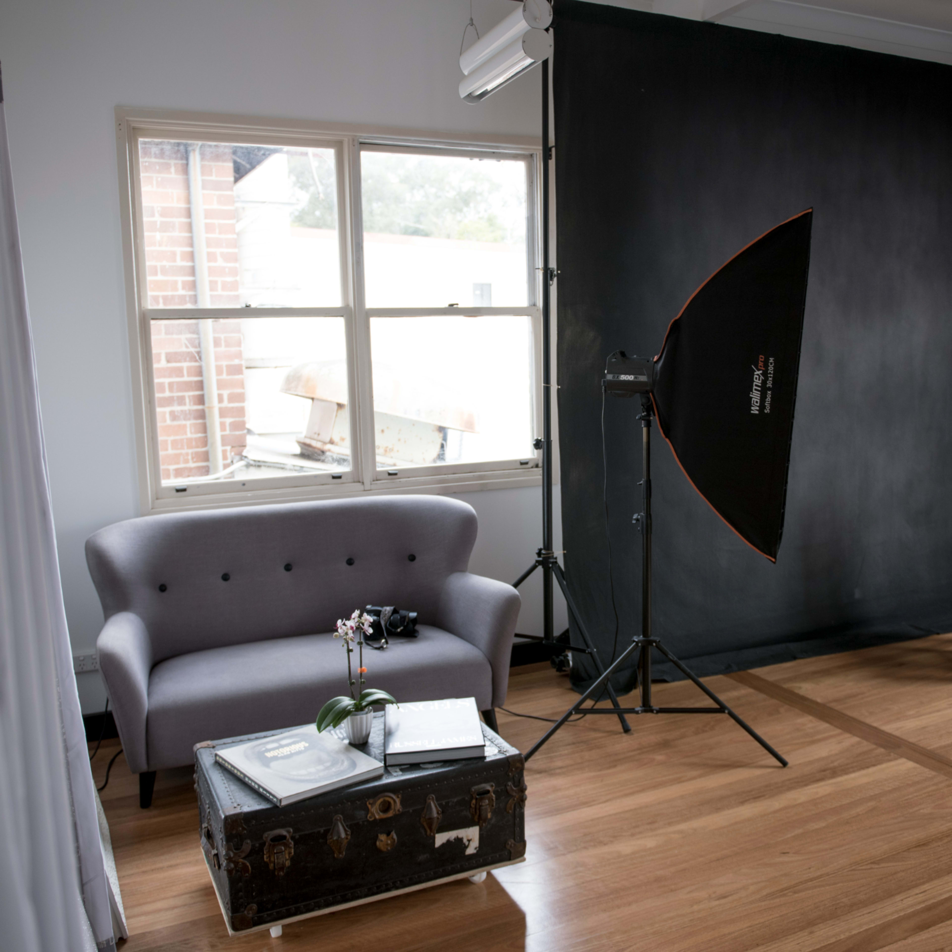 A photography studio with a gray sofa, a black backdrop, a softbox light setup, a chair, and a window allowing natural light to enter.