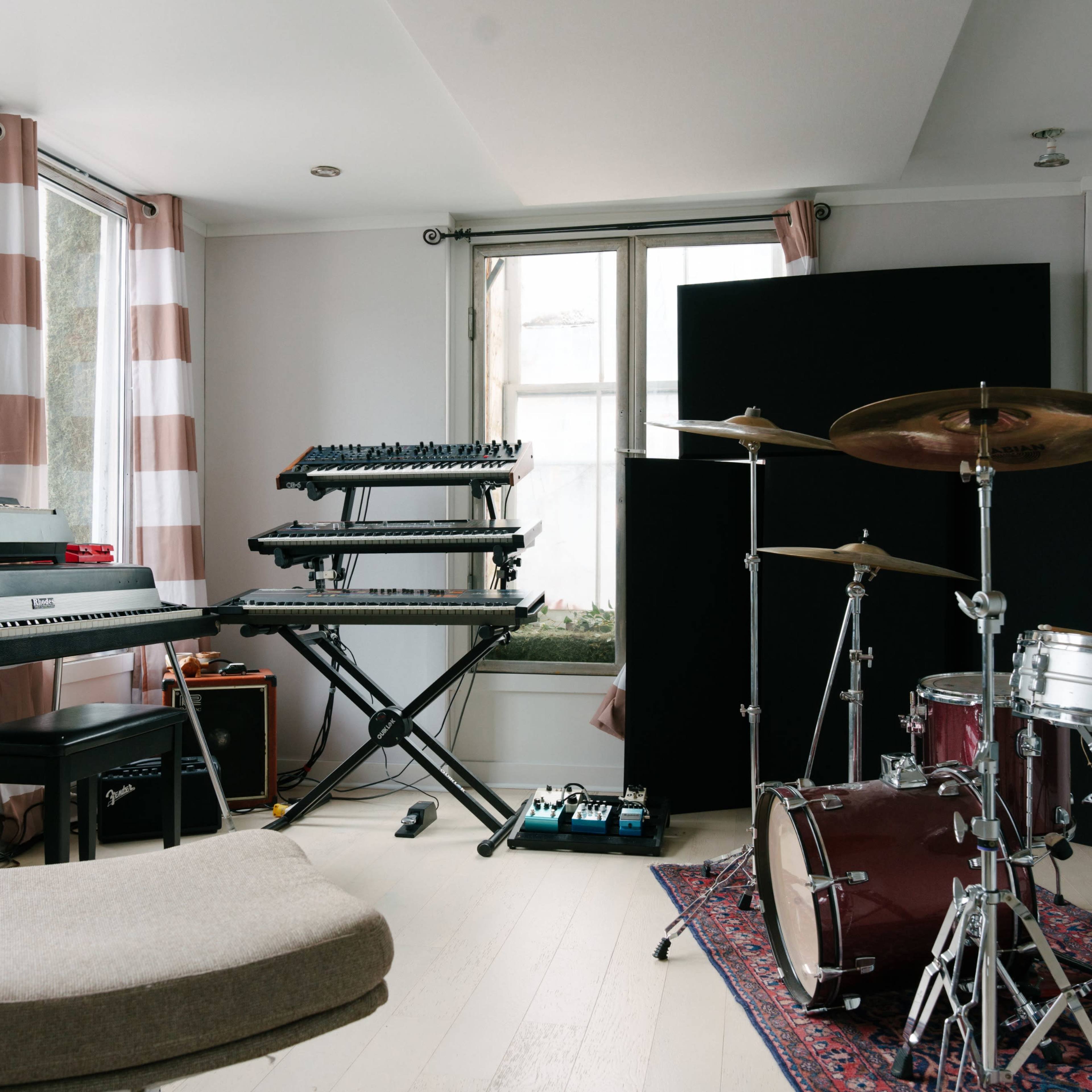 A music studio features a keyboard, synthesizer, and drum set arranged around natural light from large windows.