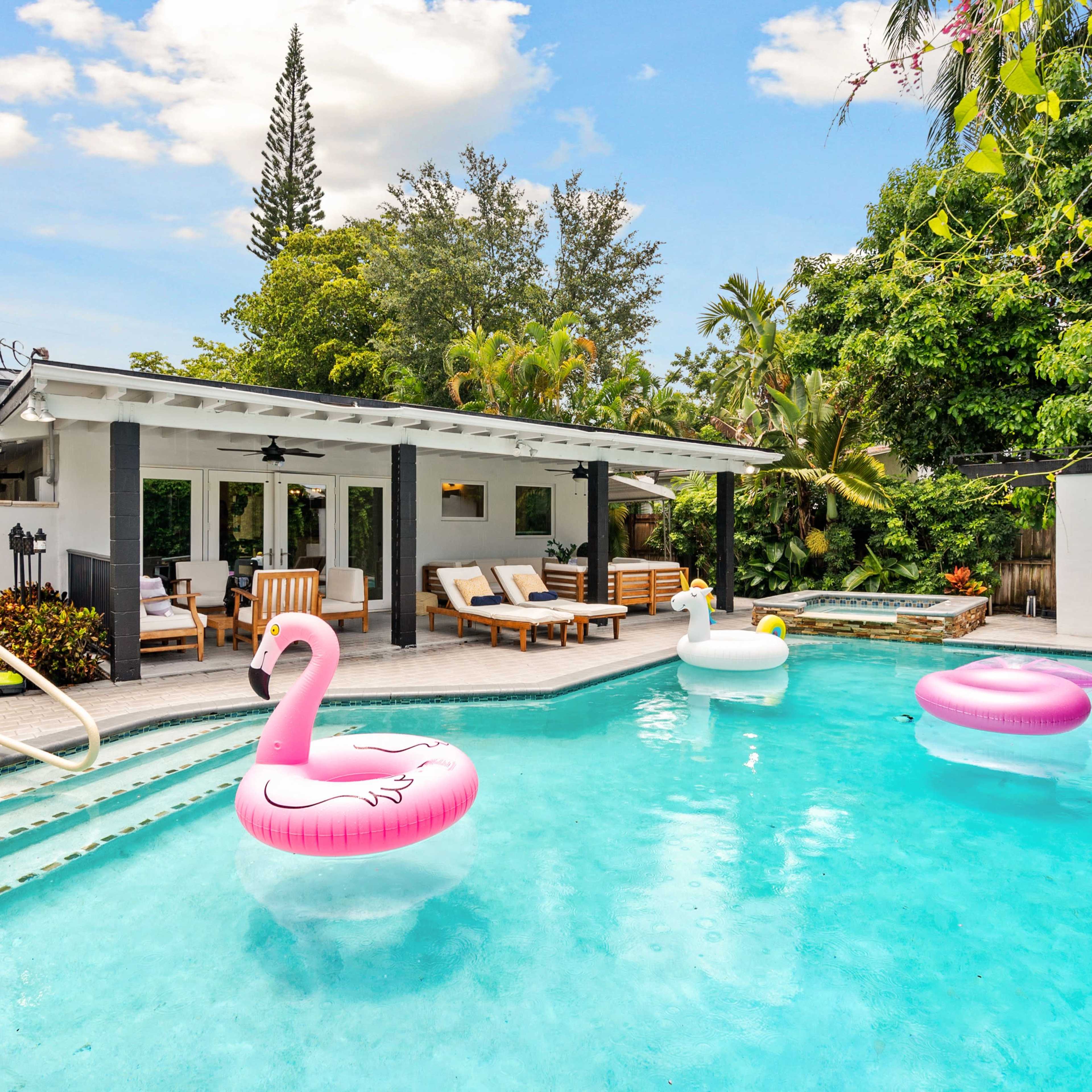 A swimming pool features pink and white inflatable floats, surrounded by lounge chairs and greenery.