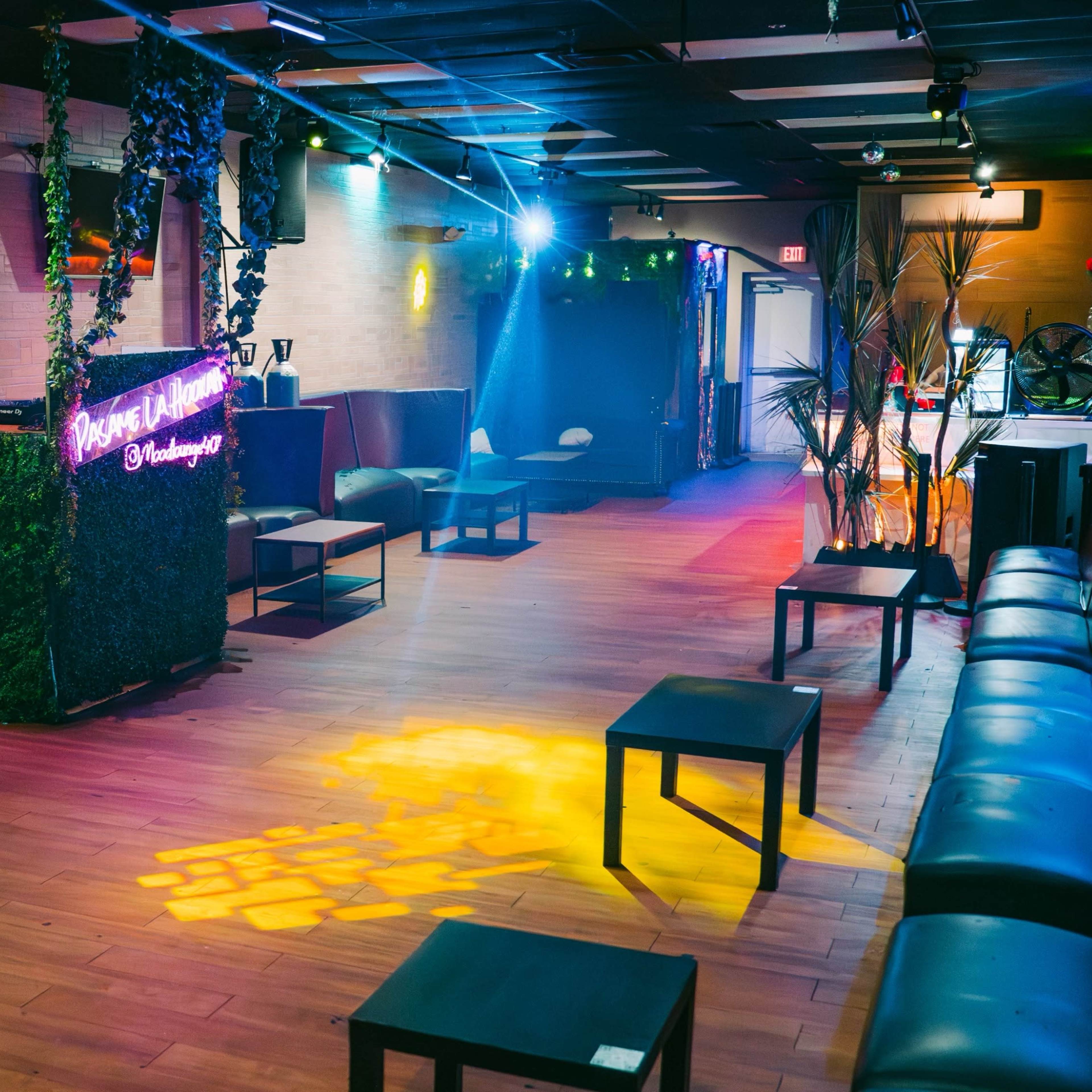An empty lounge with wooden flooring, several black tables, and a neon-lit bar area surrounded by green plants.