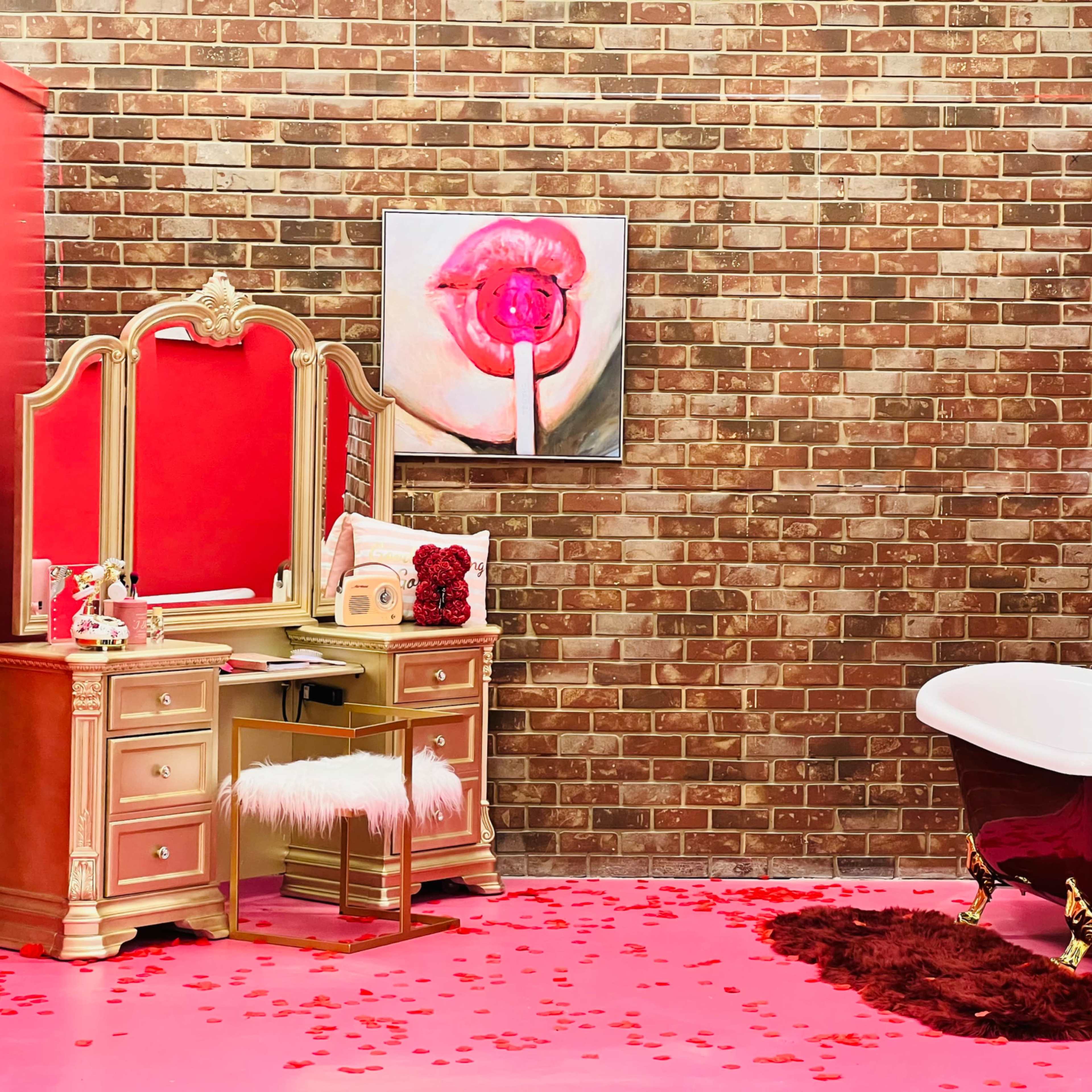 A bathroom featuring a brick wall, a red-painted space with a vintage vanity, a white freestanding bathtub, and a pink carpet scattered with rose petals.
