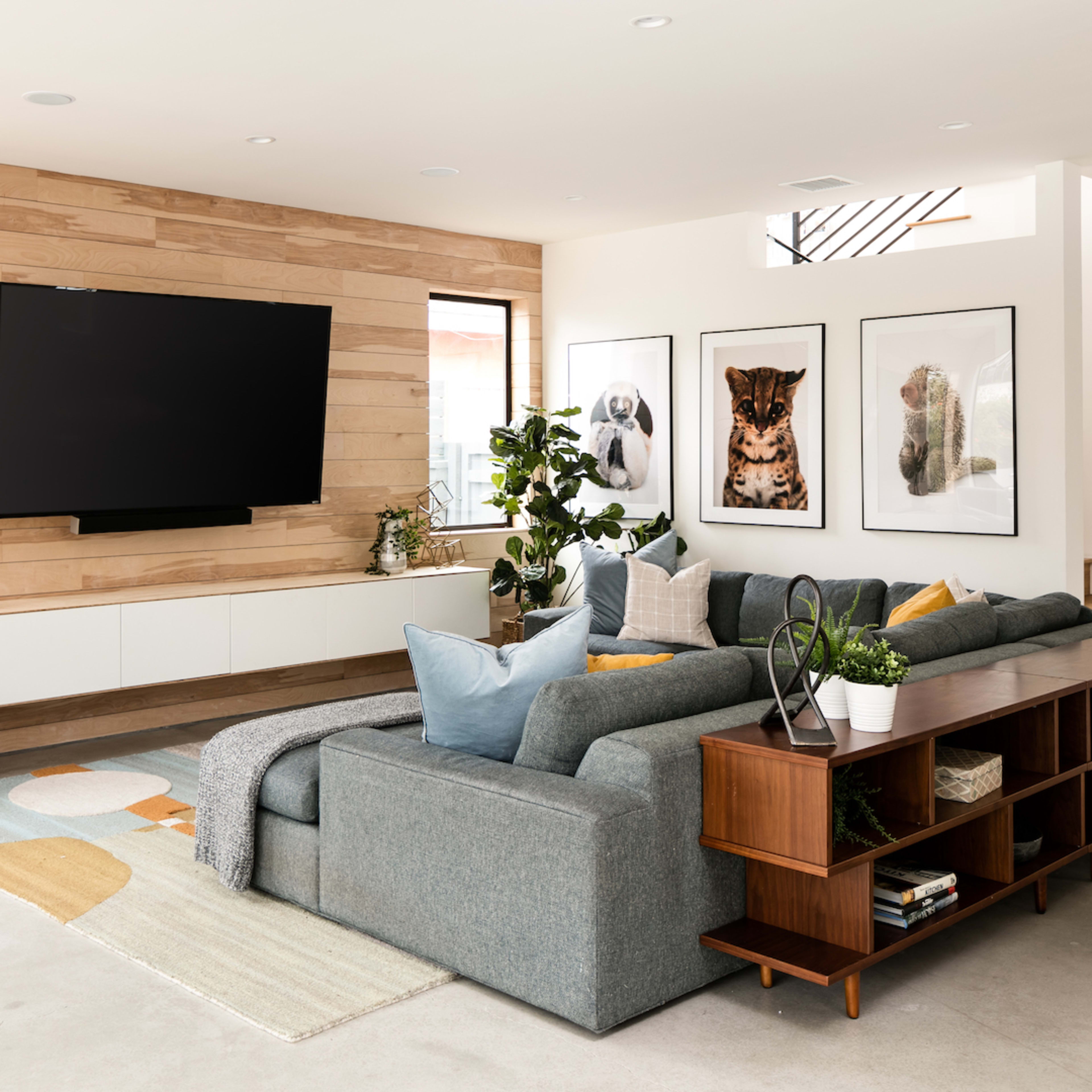 A modern living room features a large wall-mounted television, wooden accents, and a sectional sofa arranged around a patterned area rug, with artwork and plants decorating the space.