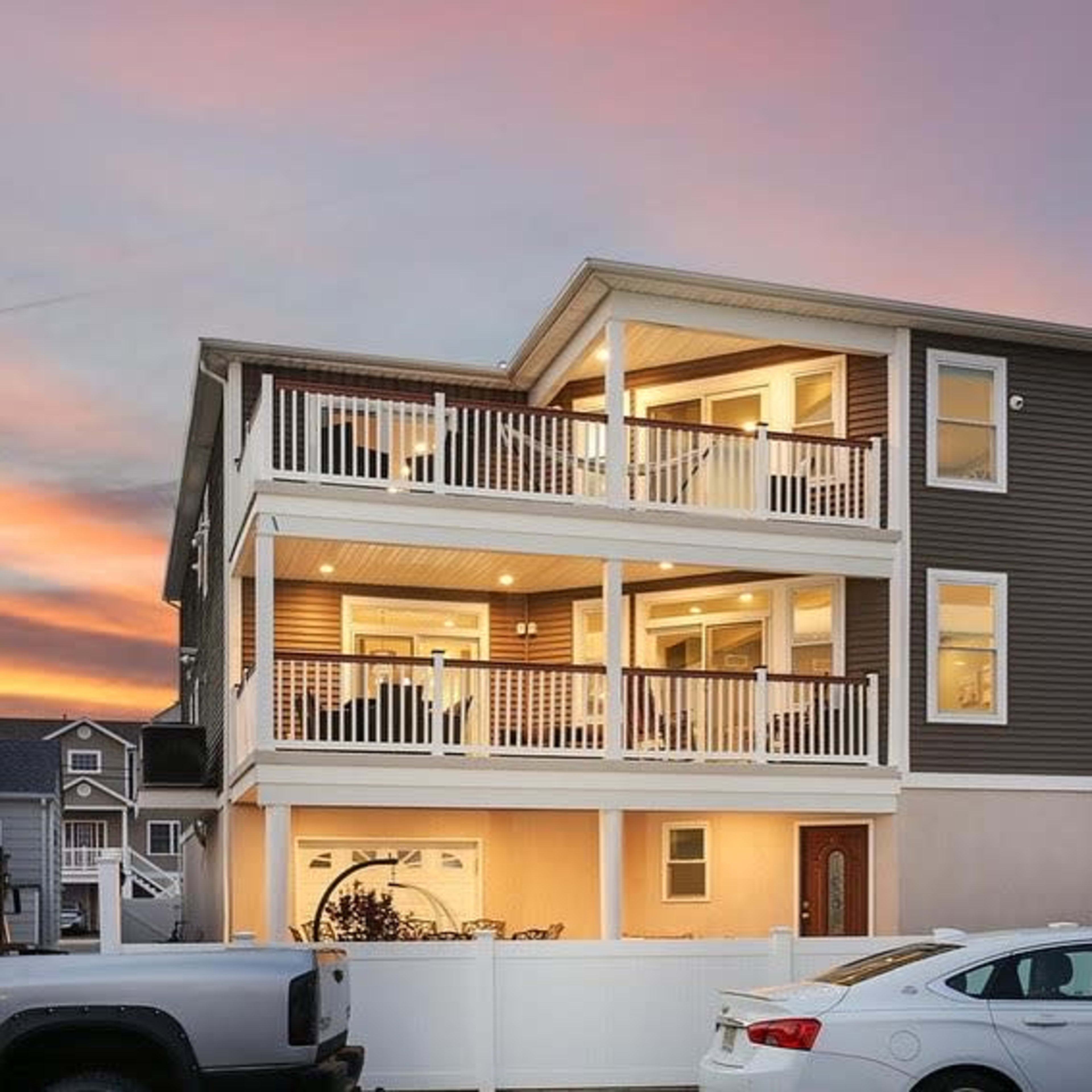 A three-story house features two balconies with white railings and large windows, illuminated at sunset, surrounded by a white fence and parked cars.