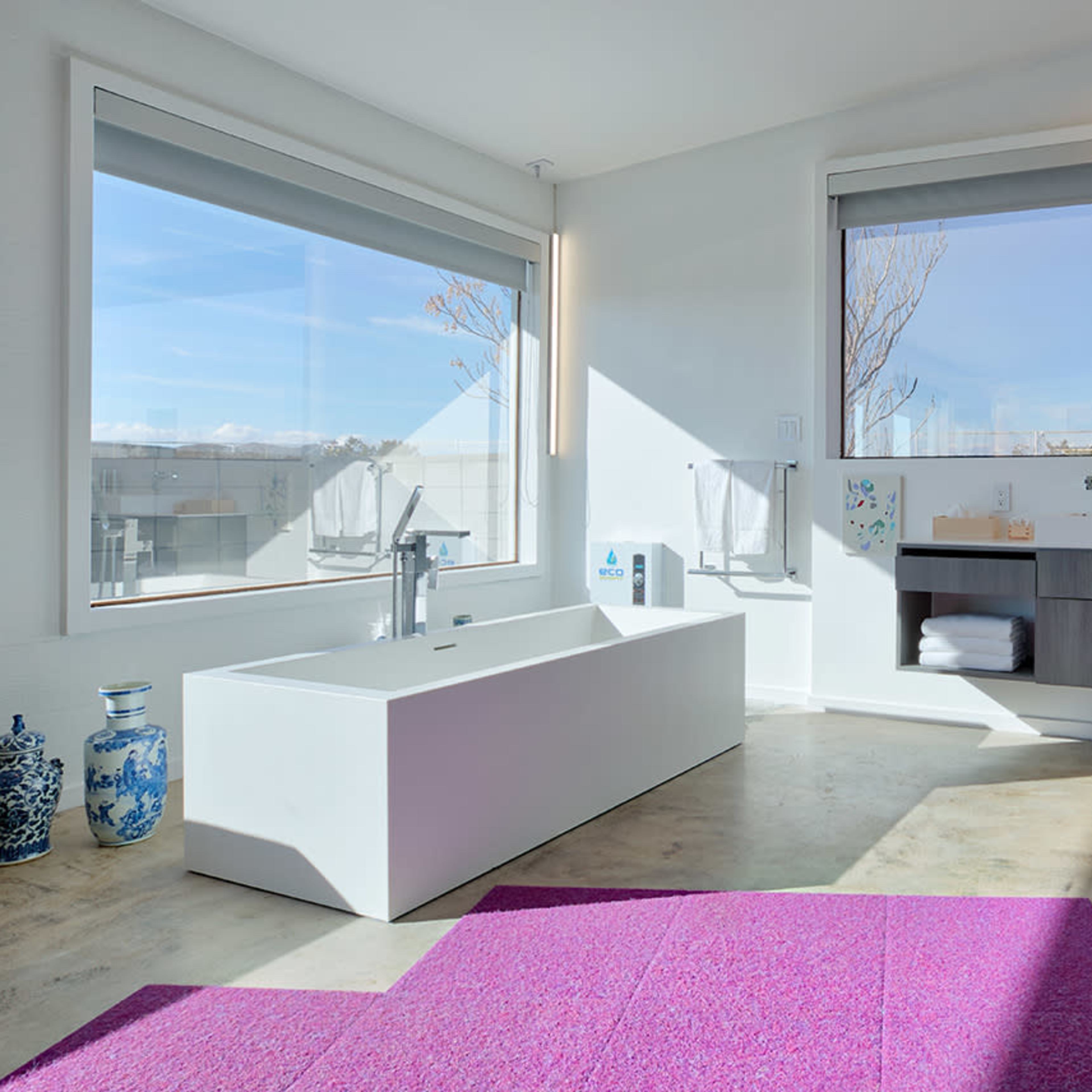 A modern bathroom features a freestanding bathtub, a wall-mounted sink with a storage unit, large windows, and a pink area rug.