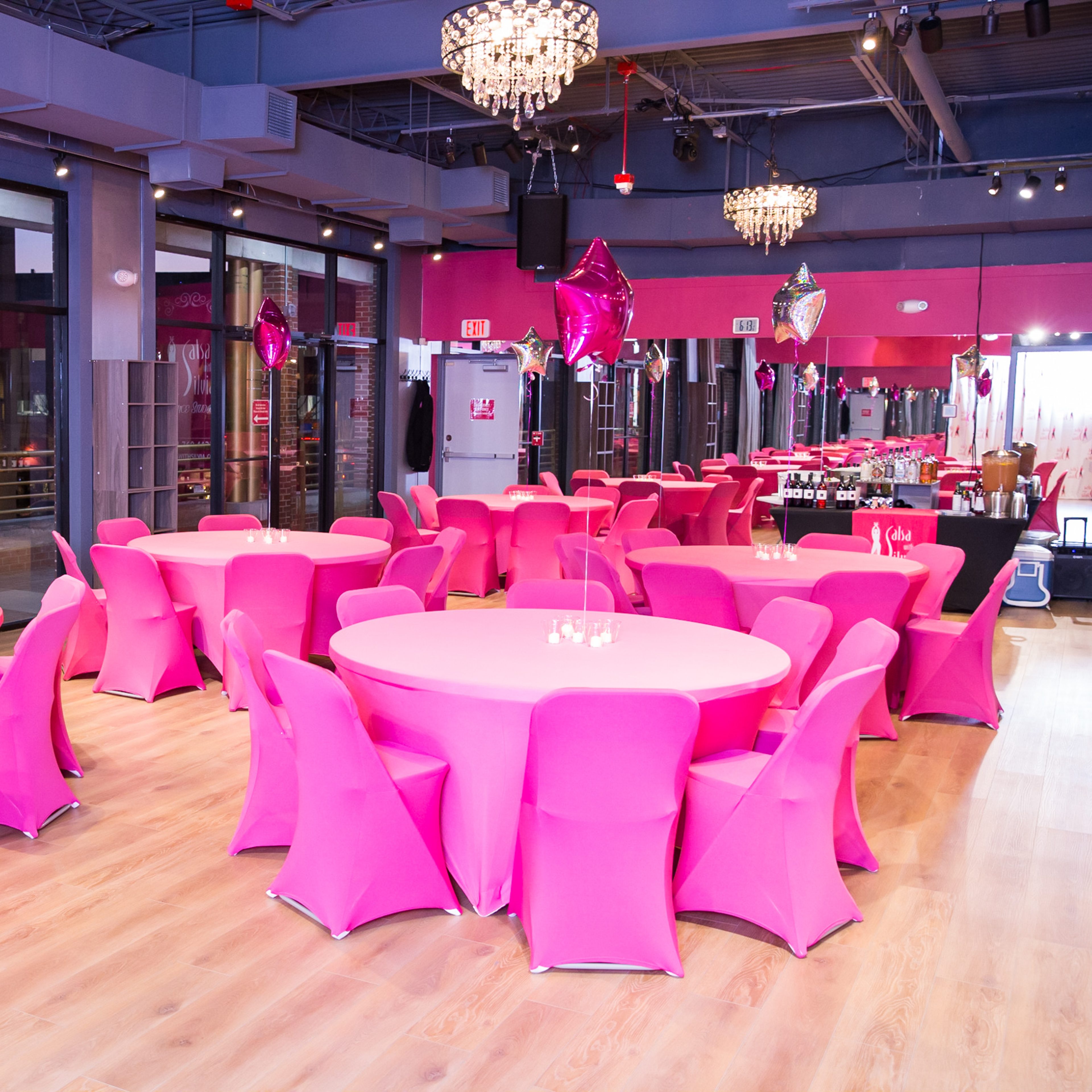 A banquet hall features round tables with pink tablecloths and chairs, illuminated by chandeliers, and decorated with balloons.