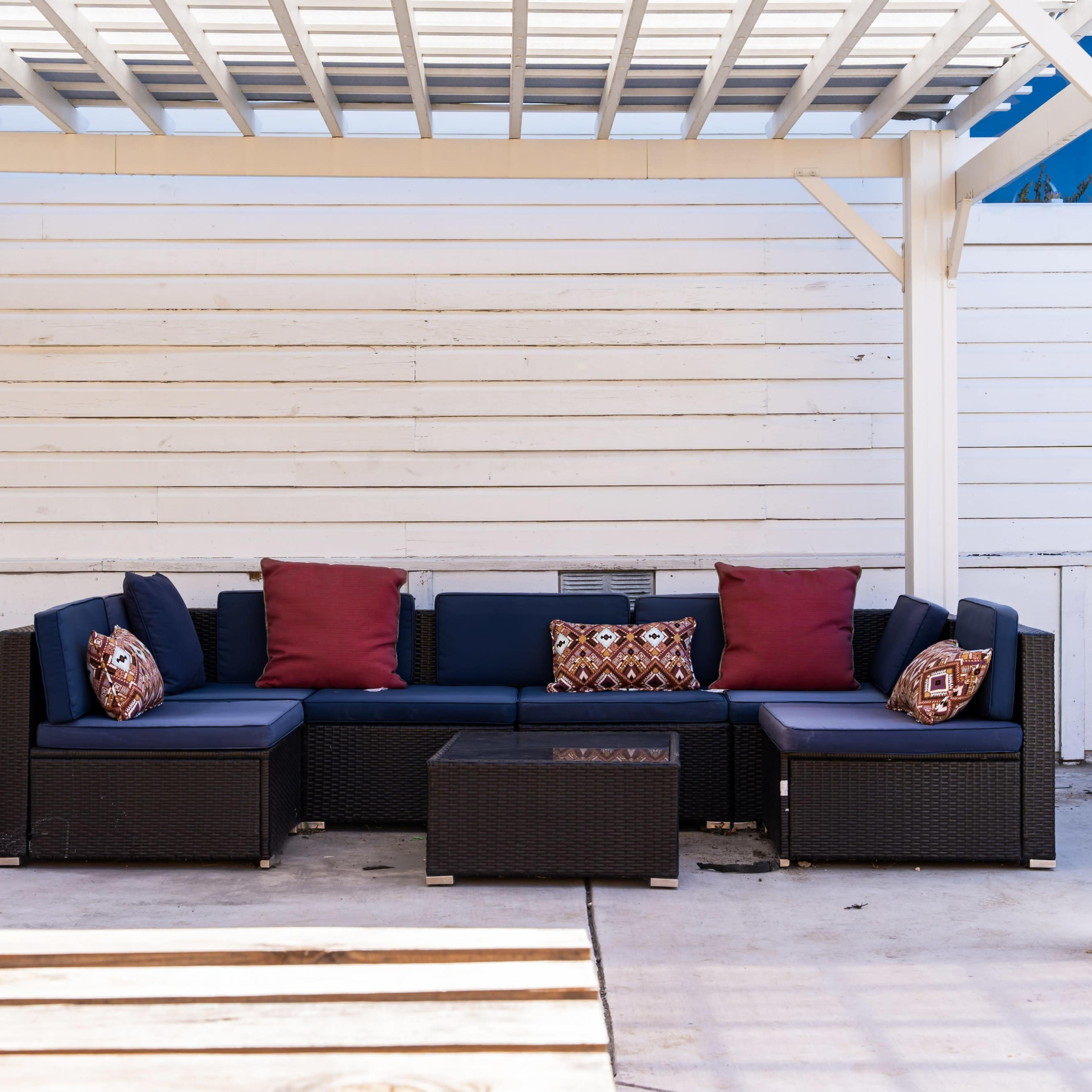 A sectional outdoor sofa with red and patterned cushions is positioned under a pergola on a concrete patio.