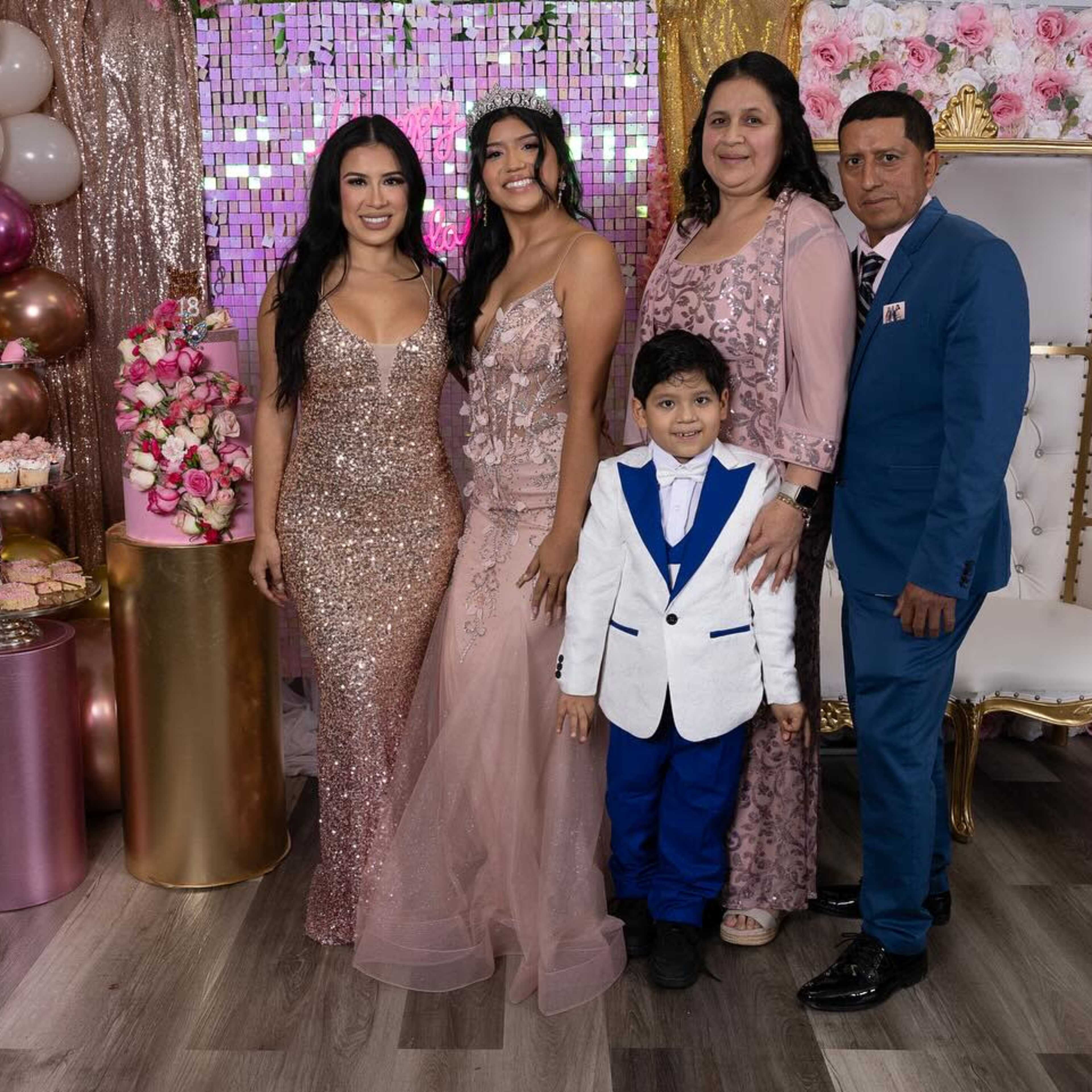 A group of five people poses together in a decorated room featuring floral backdrops and balloon arrangements for a celebration.