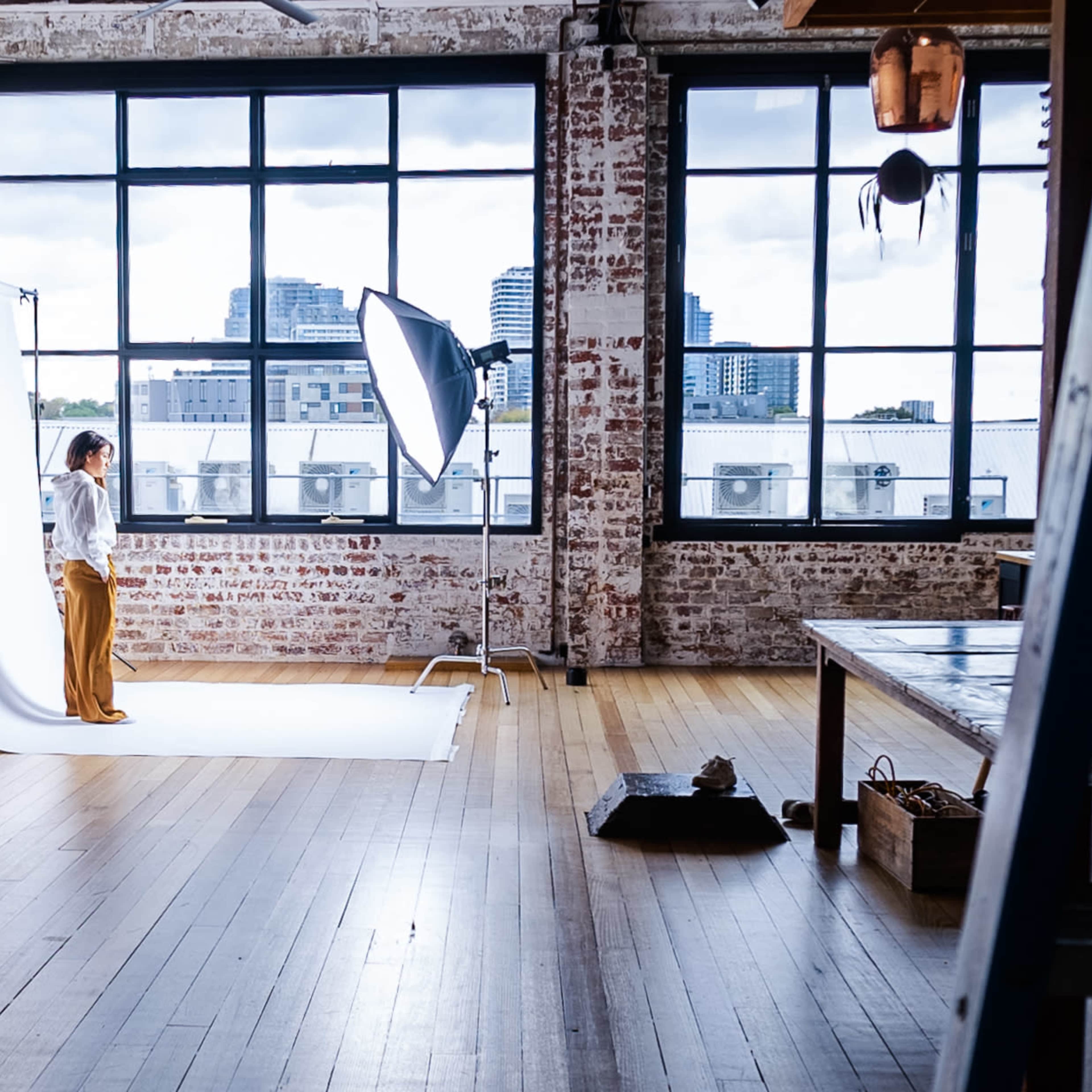 A person stands on a white backdrop in a spacious studio with large windows and exposed brick walls.
