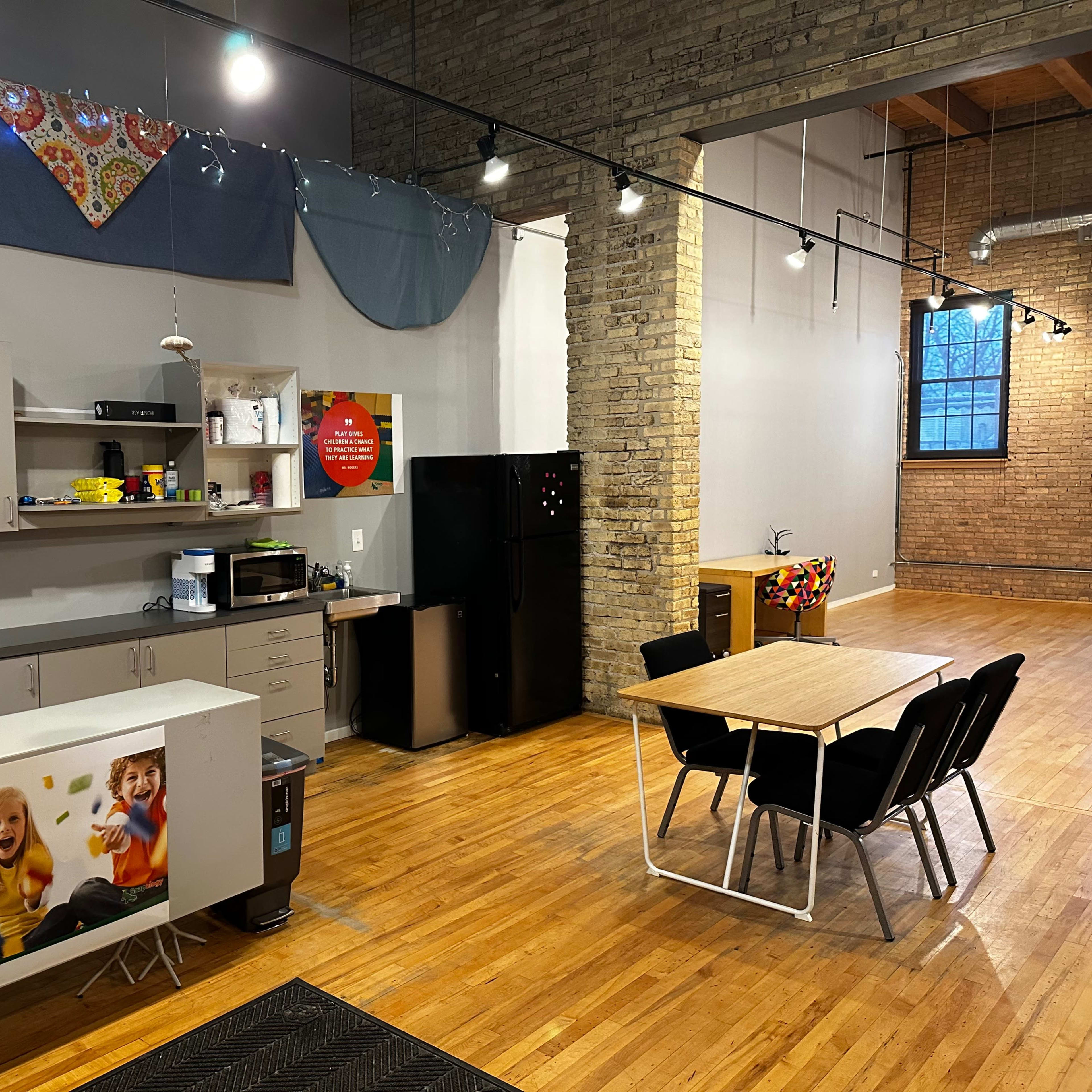 A modern kitchen area with a counter, microwave, and refrigerator, adjacent to a dining table with chairs in a room featuring exposed brick walls and wooden flooring.