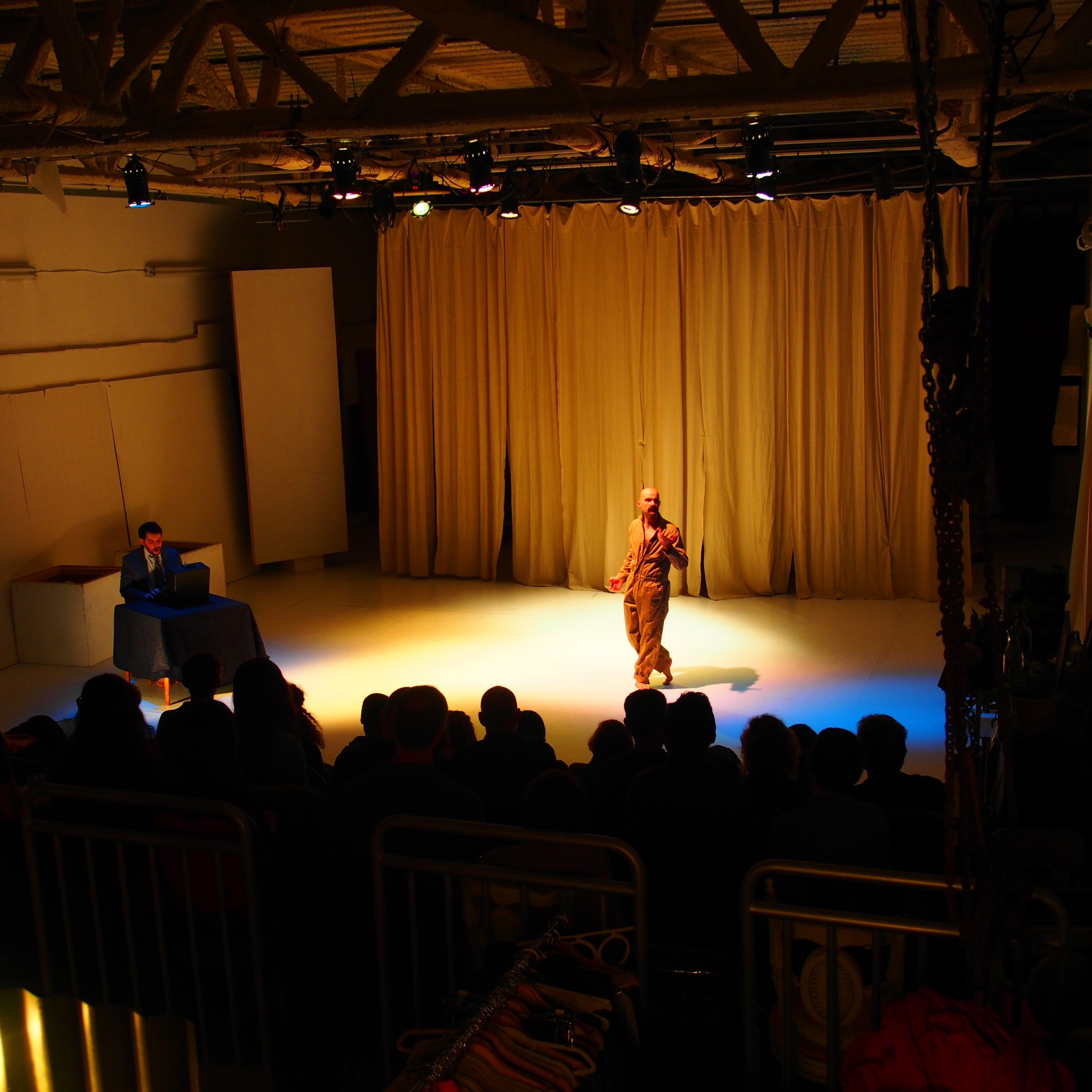 A performer stands on stage in front of a seated audience, while another individual is positioned at a table to the side, illuminated by stage lights.