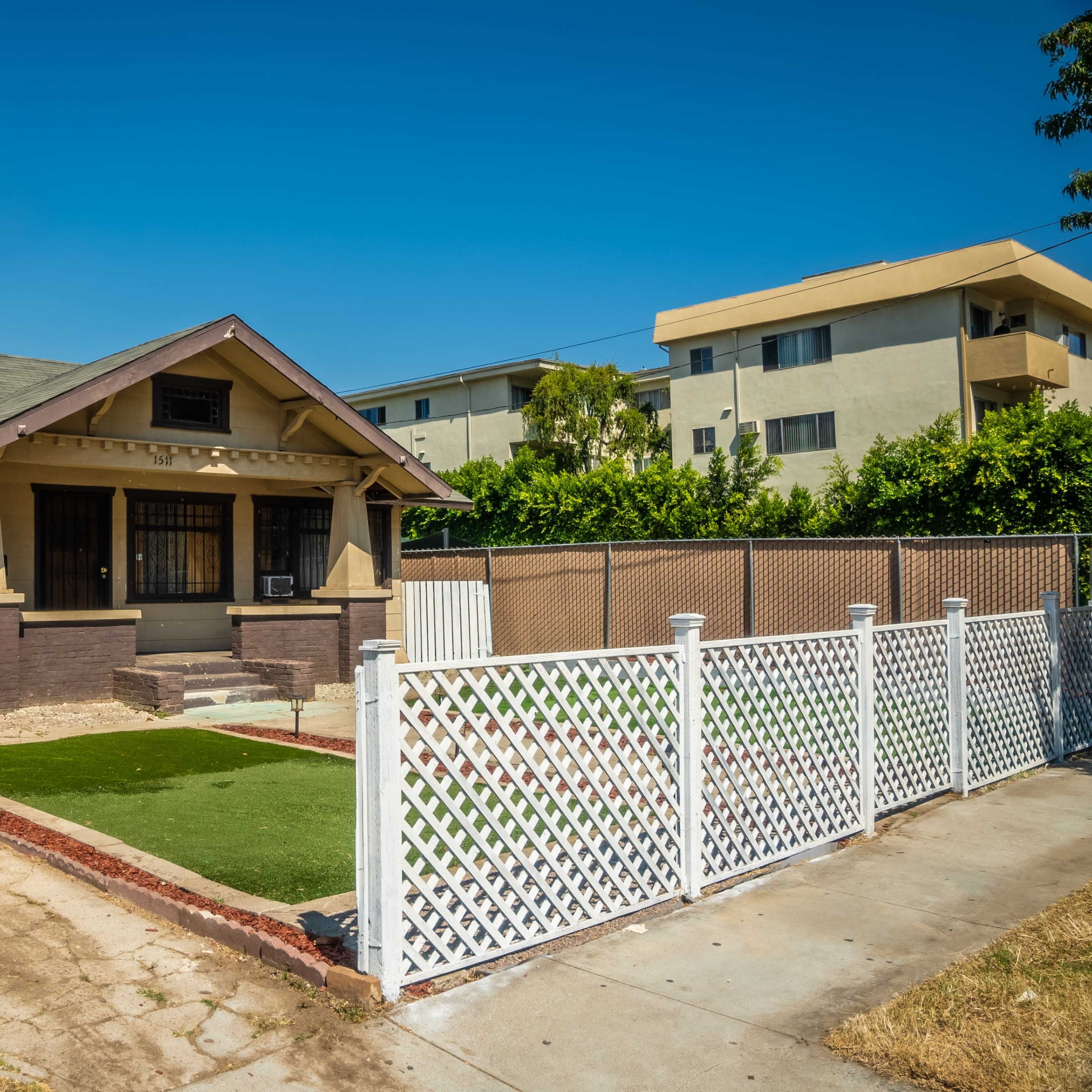 A single-story bungalow with a front yard enclosed by a white lattice fence, situated beside a multi-story apartment building.