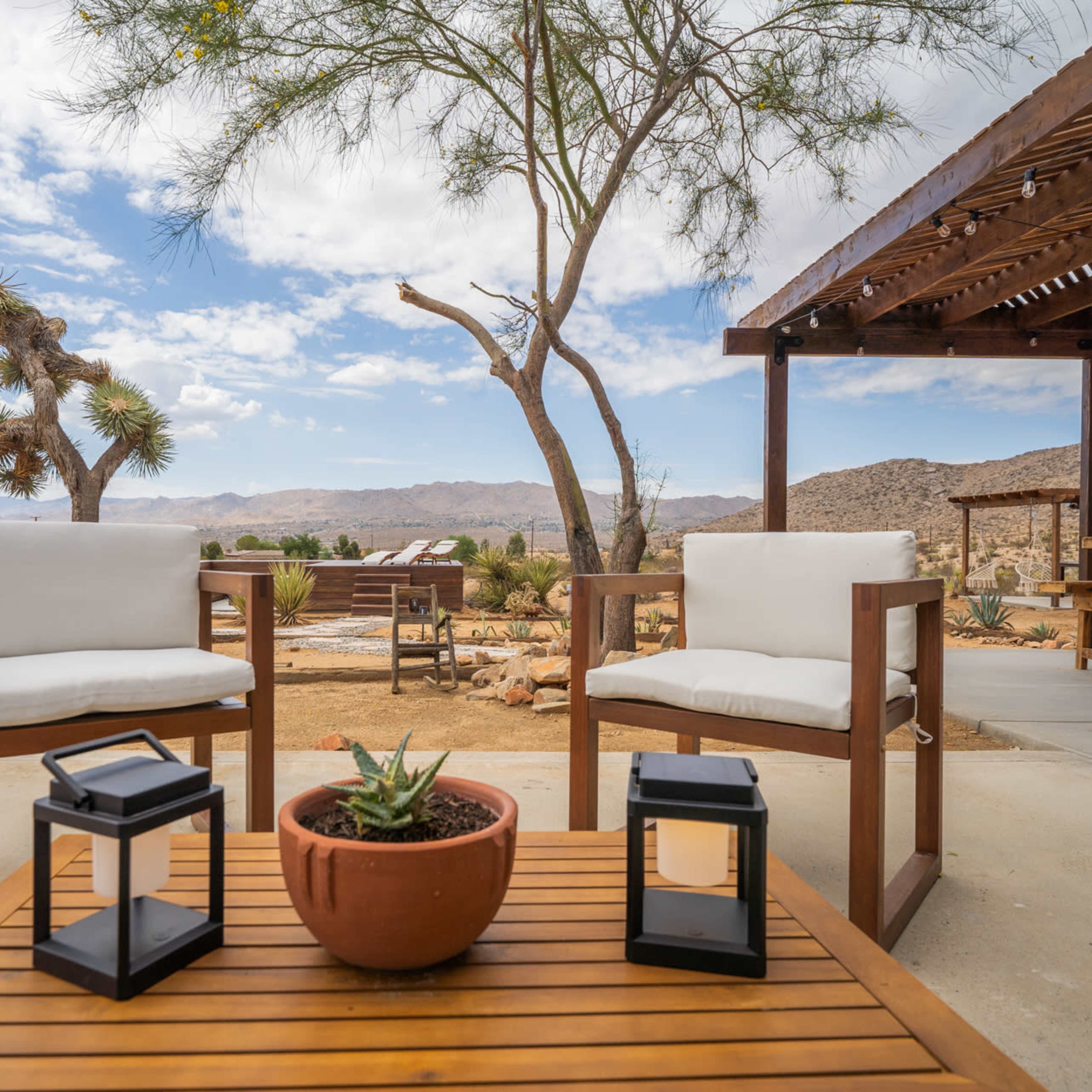 A desert patio with two white chairs, a wooden table, and a potted plant, surrounded by mountains and a cloudy sky.