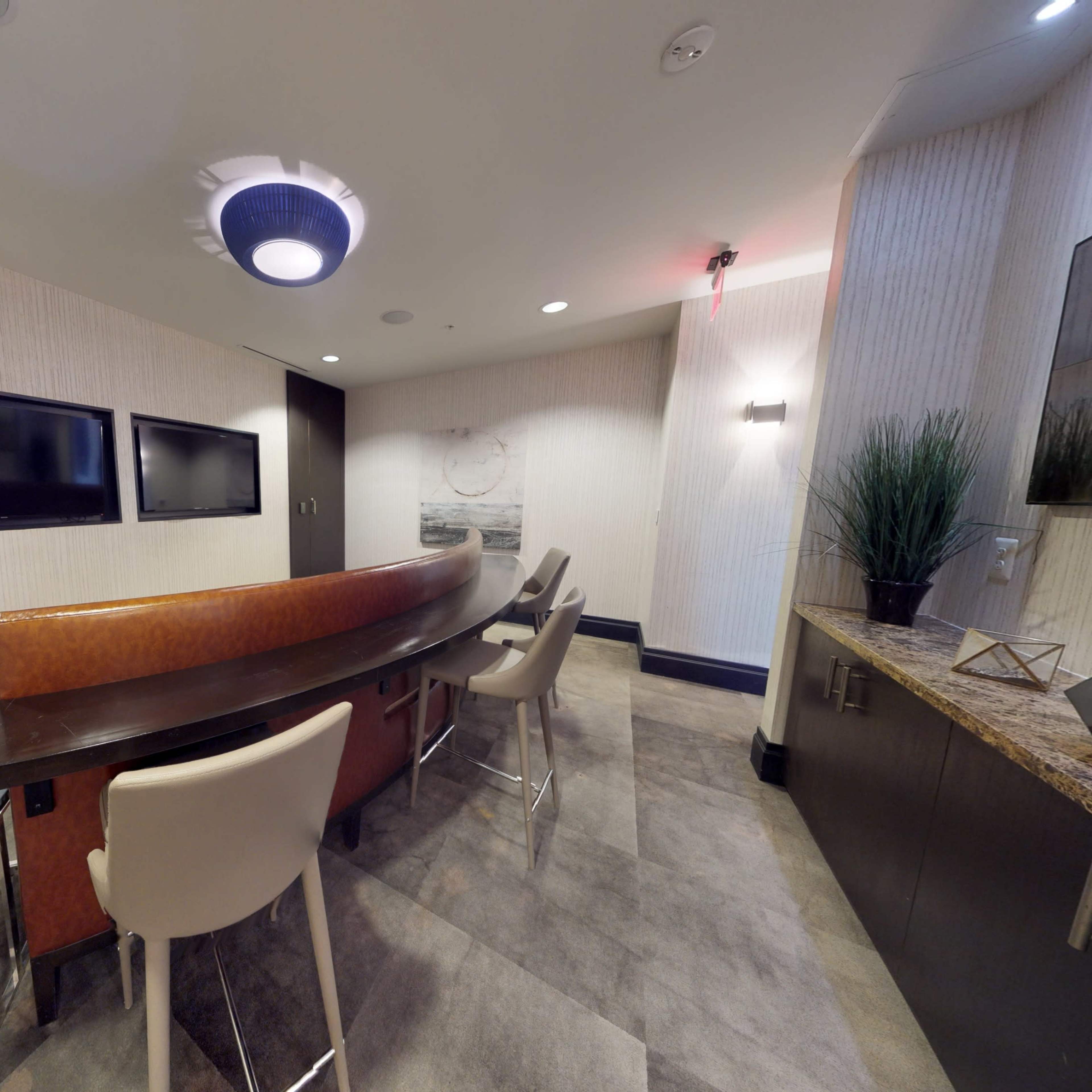 A modern, small meeting room featuring a curved wooden table, several chairs, and two wall-mounted screens.