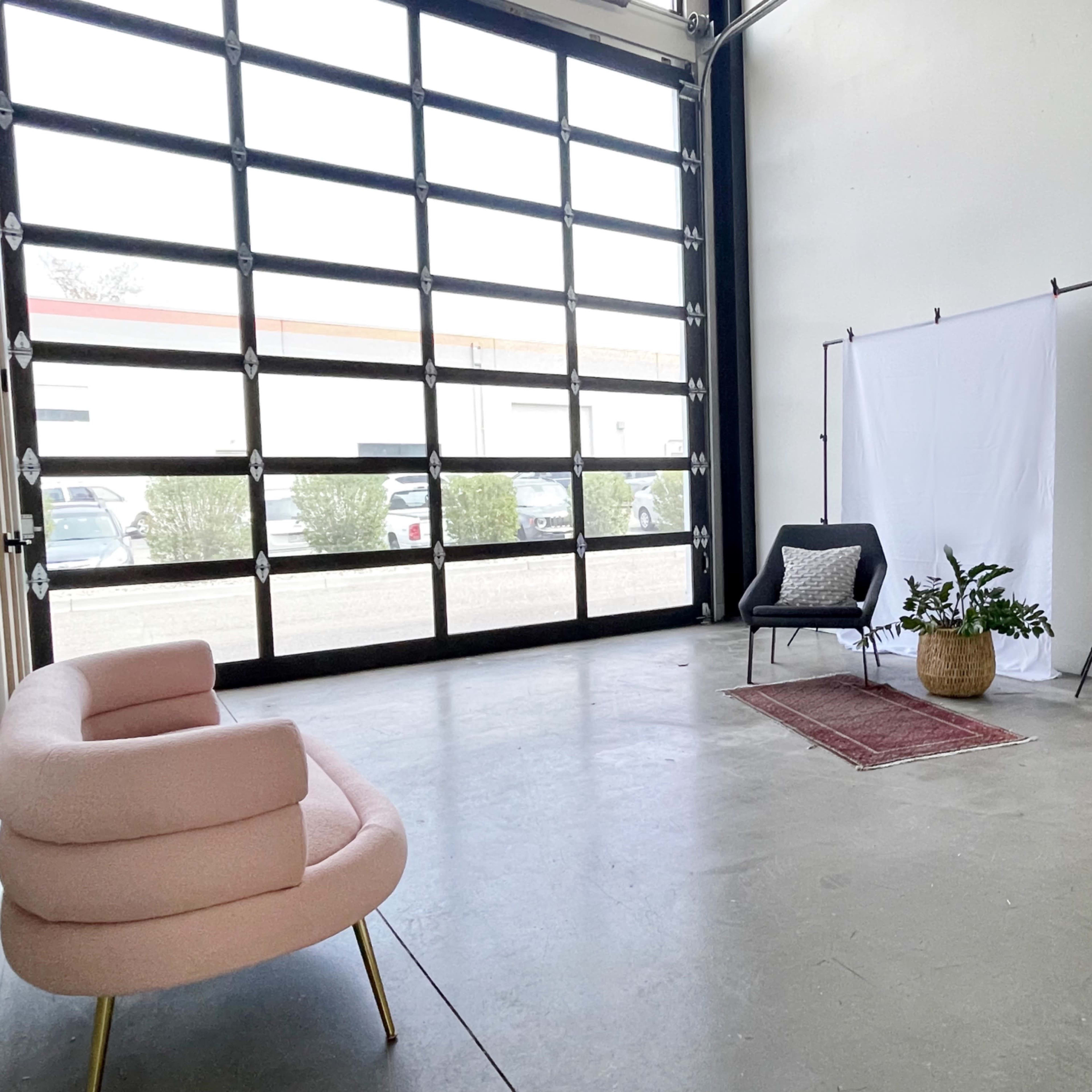 A spacious studio features a pink sofa, a white backdrop supported by stands, and two lighting setups next to a large glass garage door.