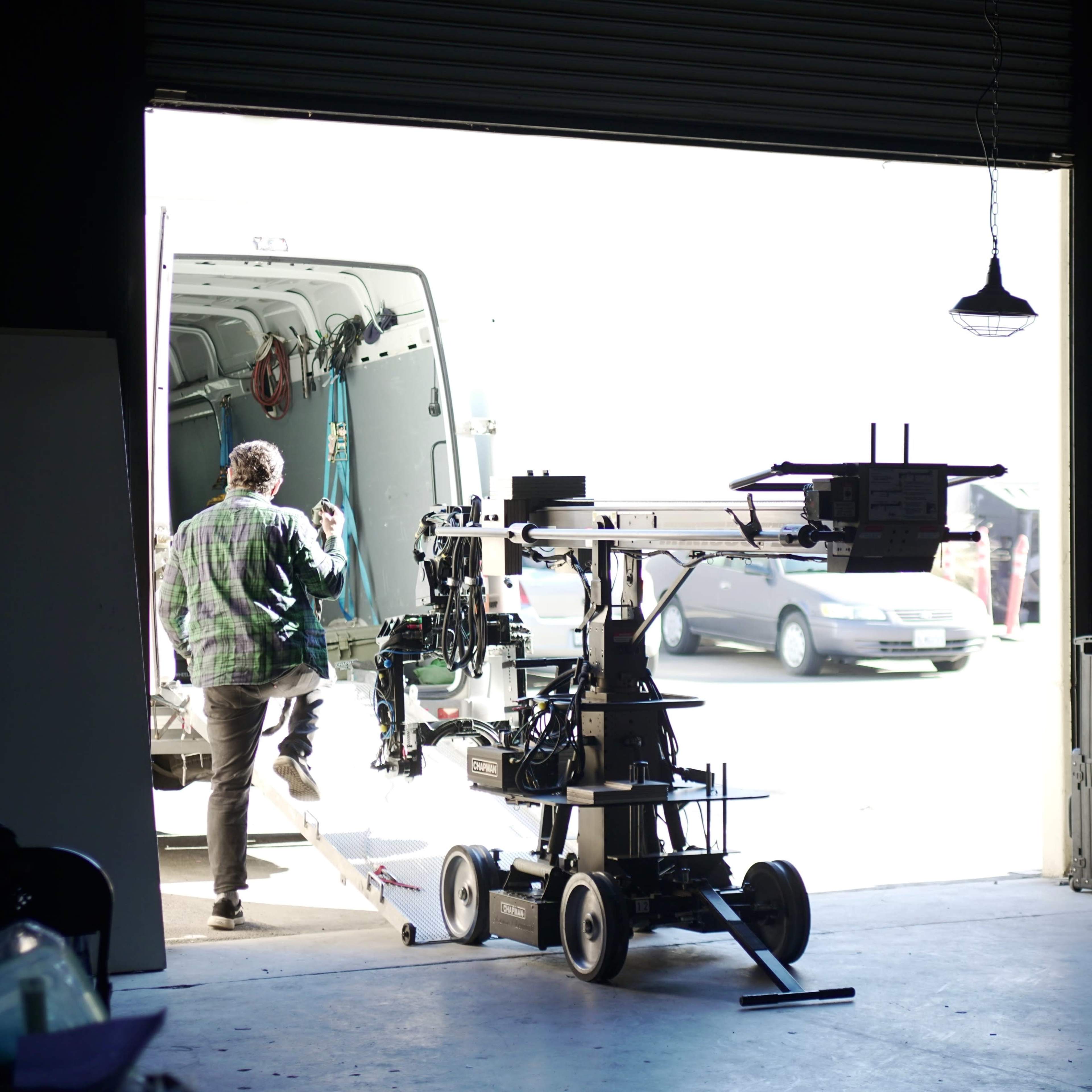 A person in a plaid shirt is unloading film equipment from a van in a well-lit warehouse.