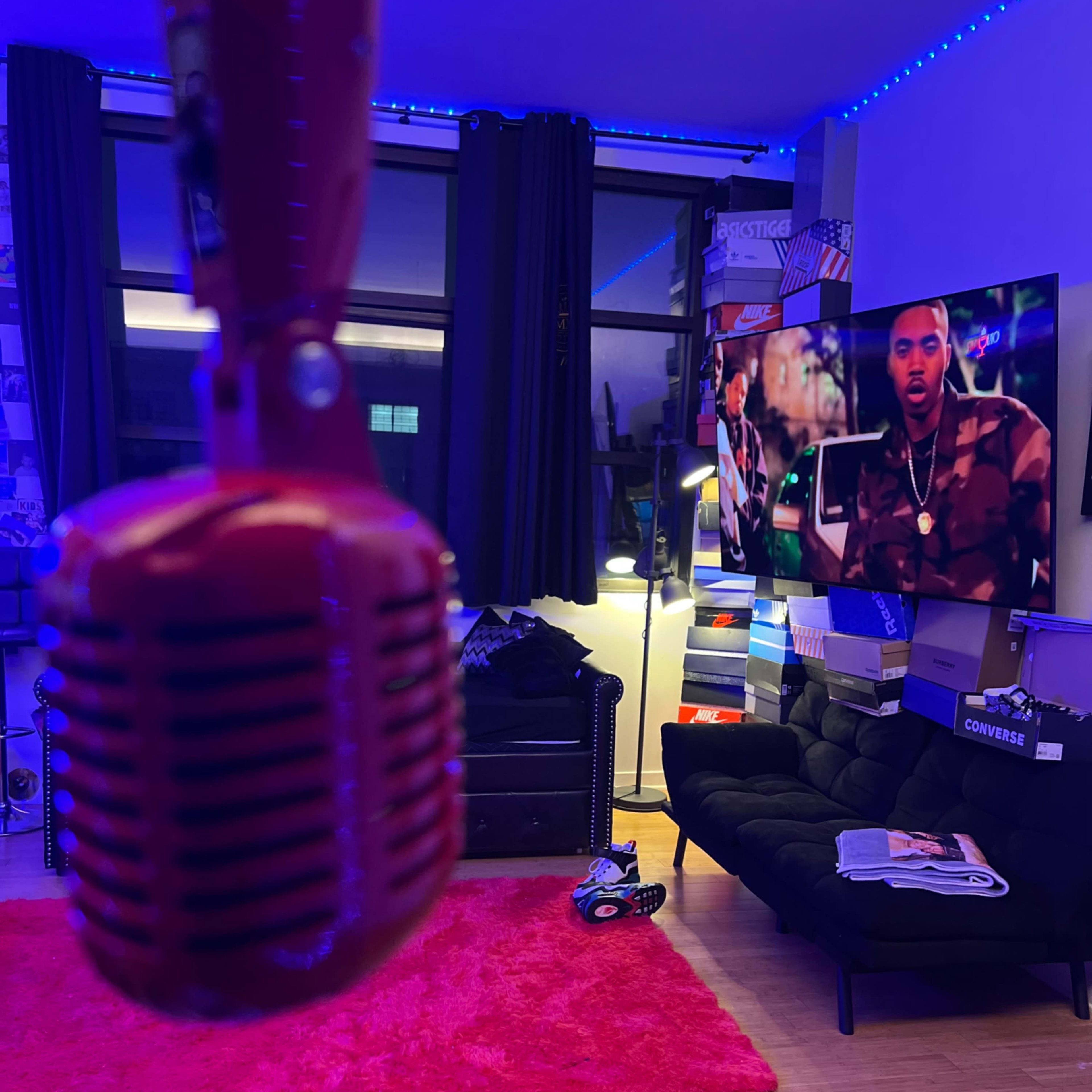 A vintage red microphone hangs in the foreground with a large television displaying a man in a camouflage jacket in a brightly lit room.