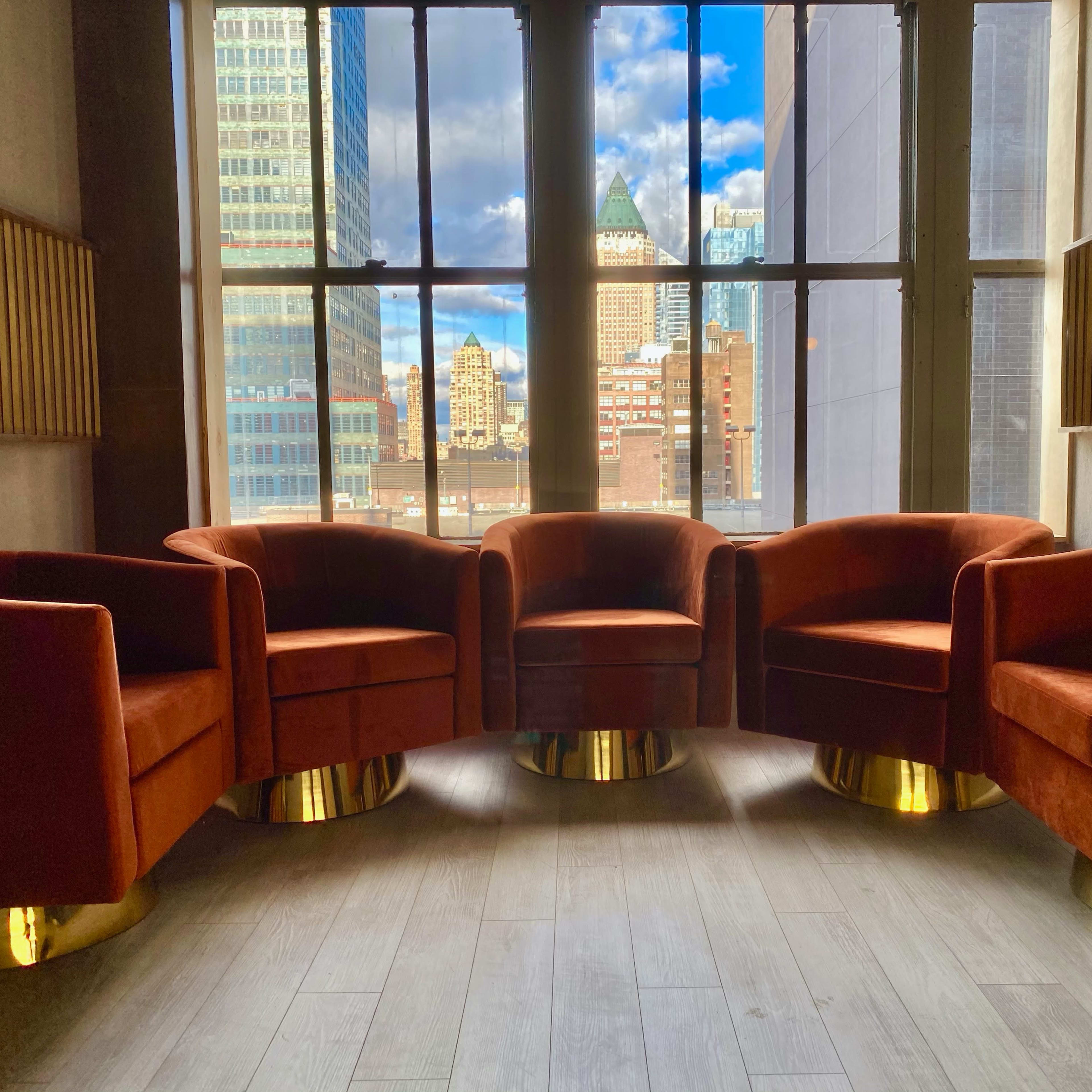 Four orange velvet chairs with gold bases are arranged in a circle near large windows, overlooking a city skyline.