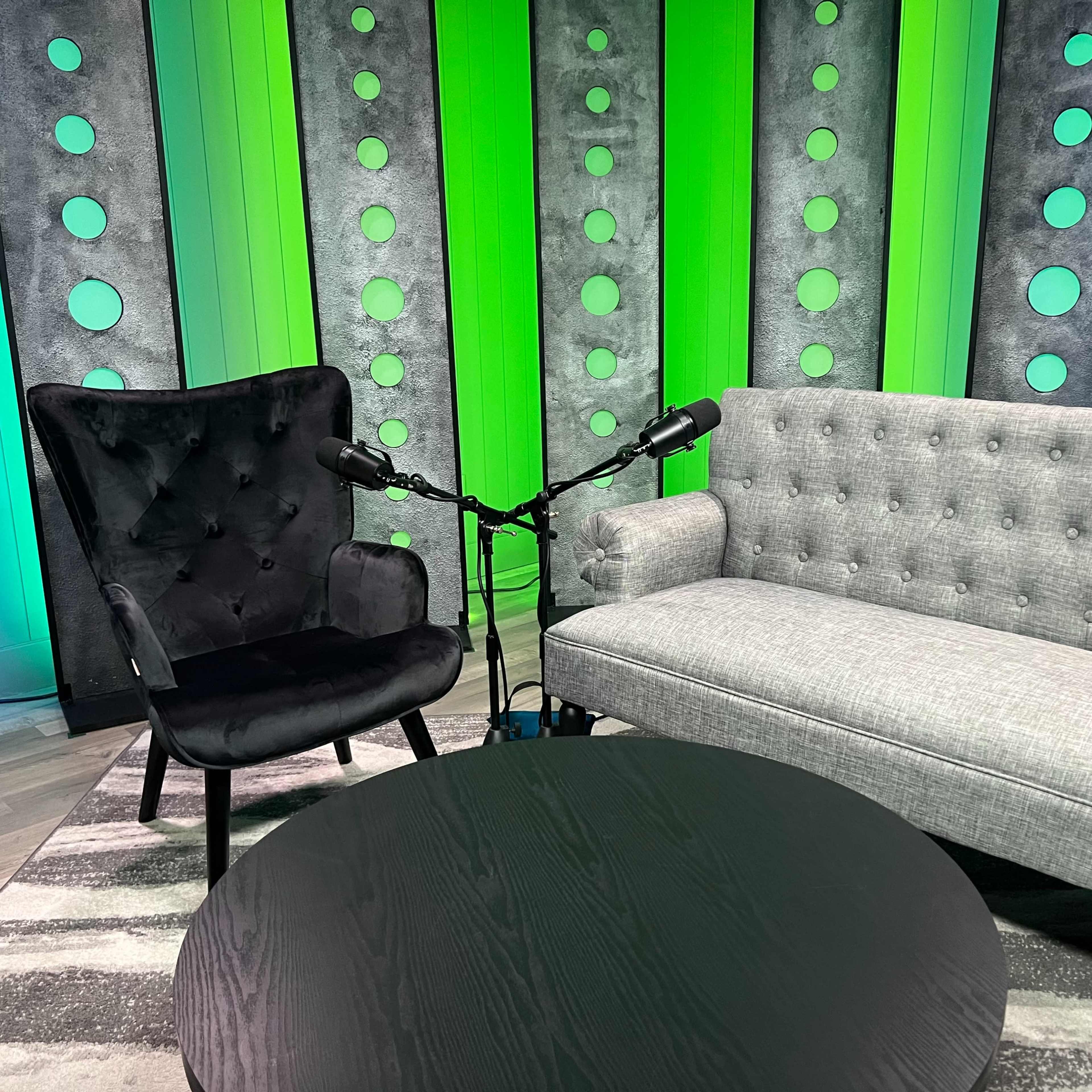 A modern interview setup featuring a black armchair and a gray tufted loveseat, with two microphones positioned on a round black coffee table, against a backdrop of green and gray panels with circular cutouts.