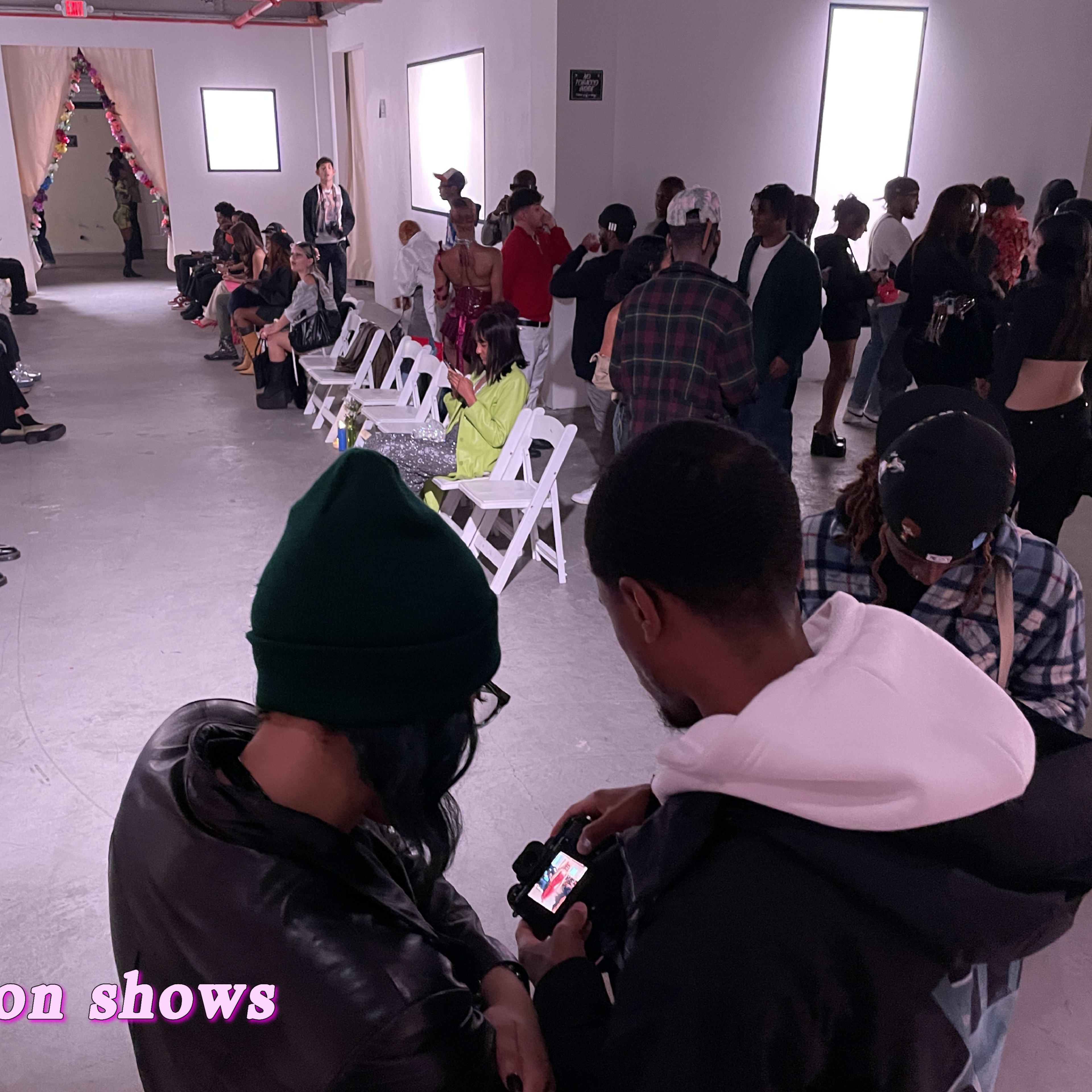 A spacious room with white walls filled with people seated and standing, preparing for a fashion show.