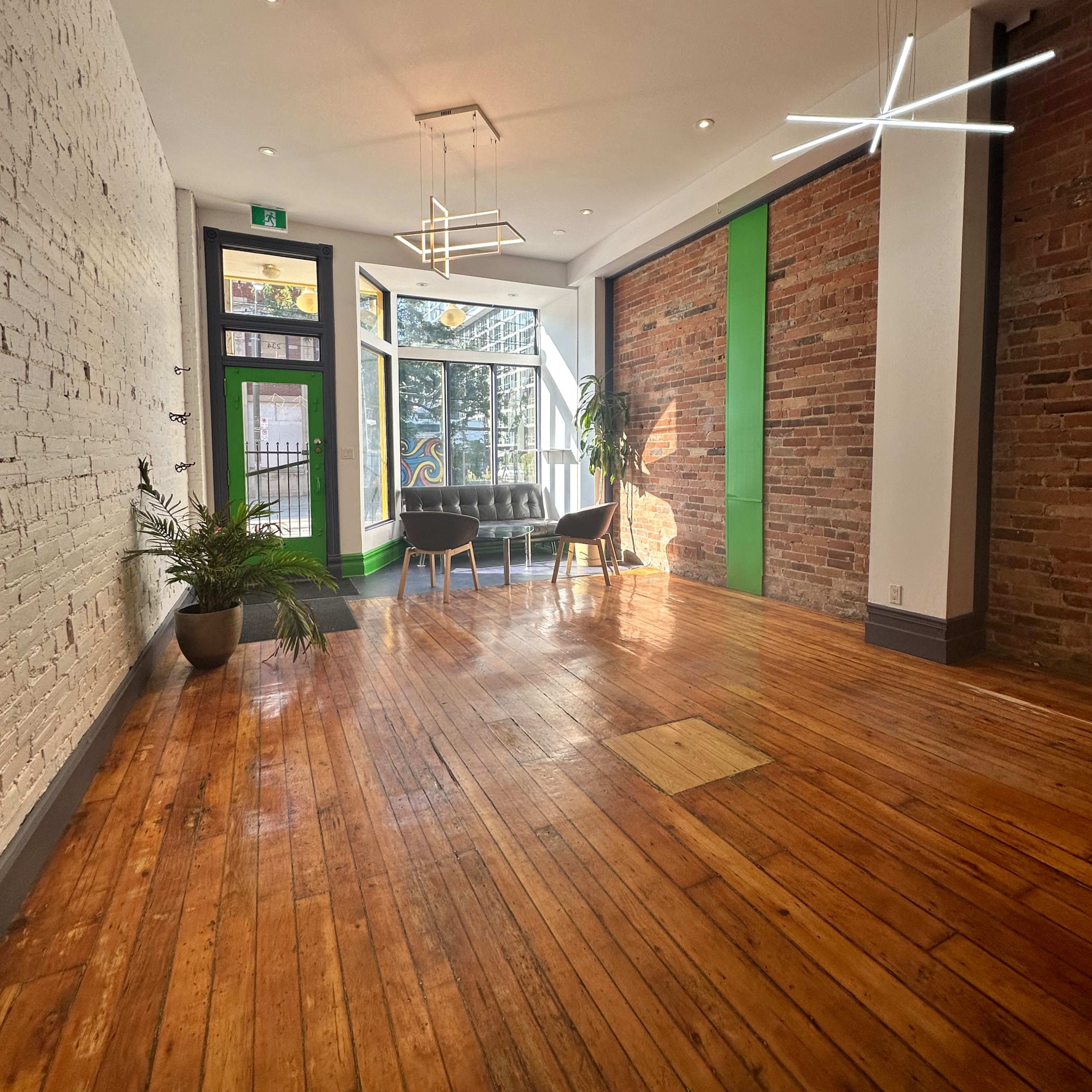 A spacious room with wooden floors, a combination of exposed brick and white walls, modern lighting fixtures, and a seating area near large windows.