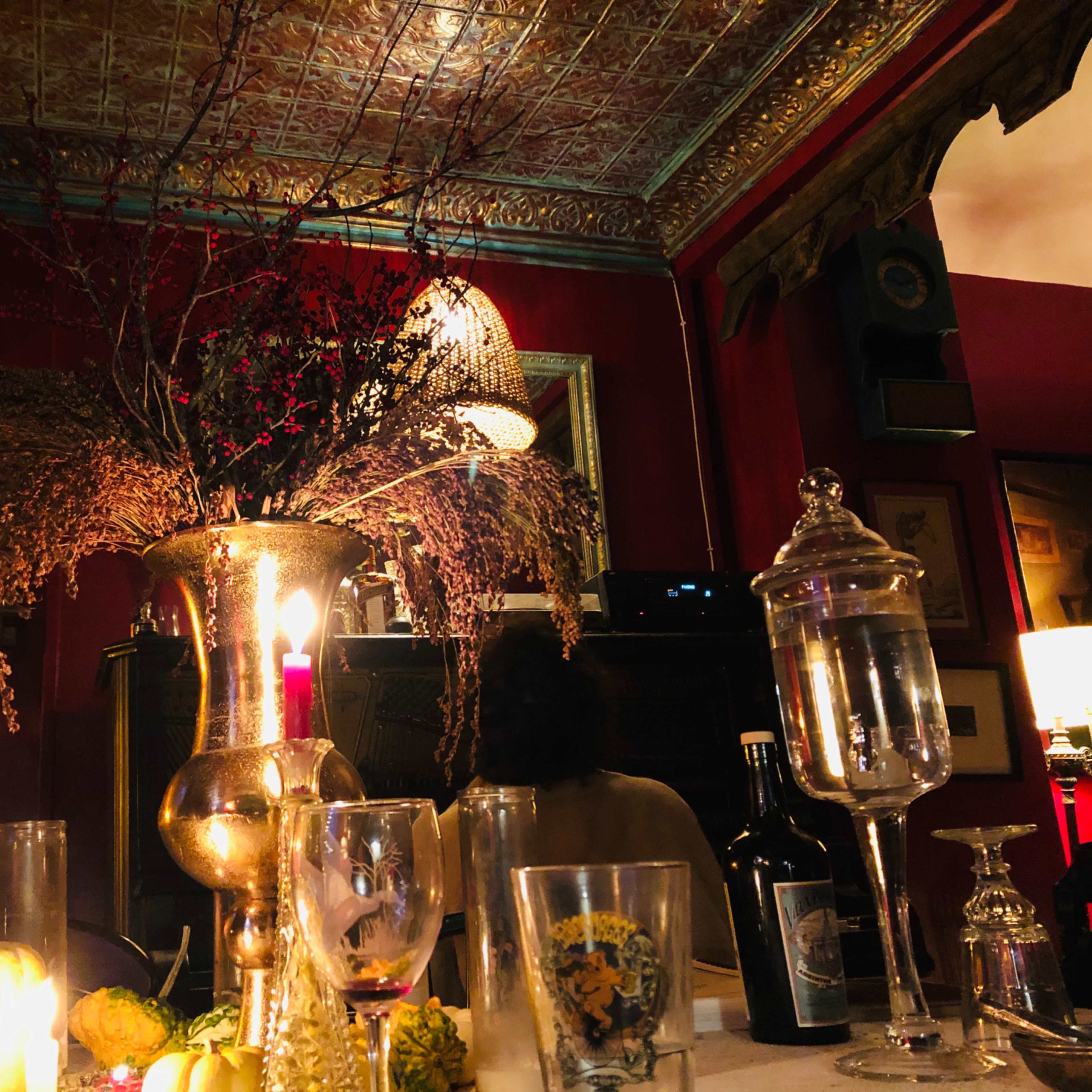 A dimly lit dining table is adorned with a large floral arrangement in a metallic vase, surrounded by various bottles, glasses, and decorative items against a richly colored wall.