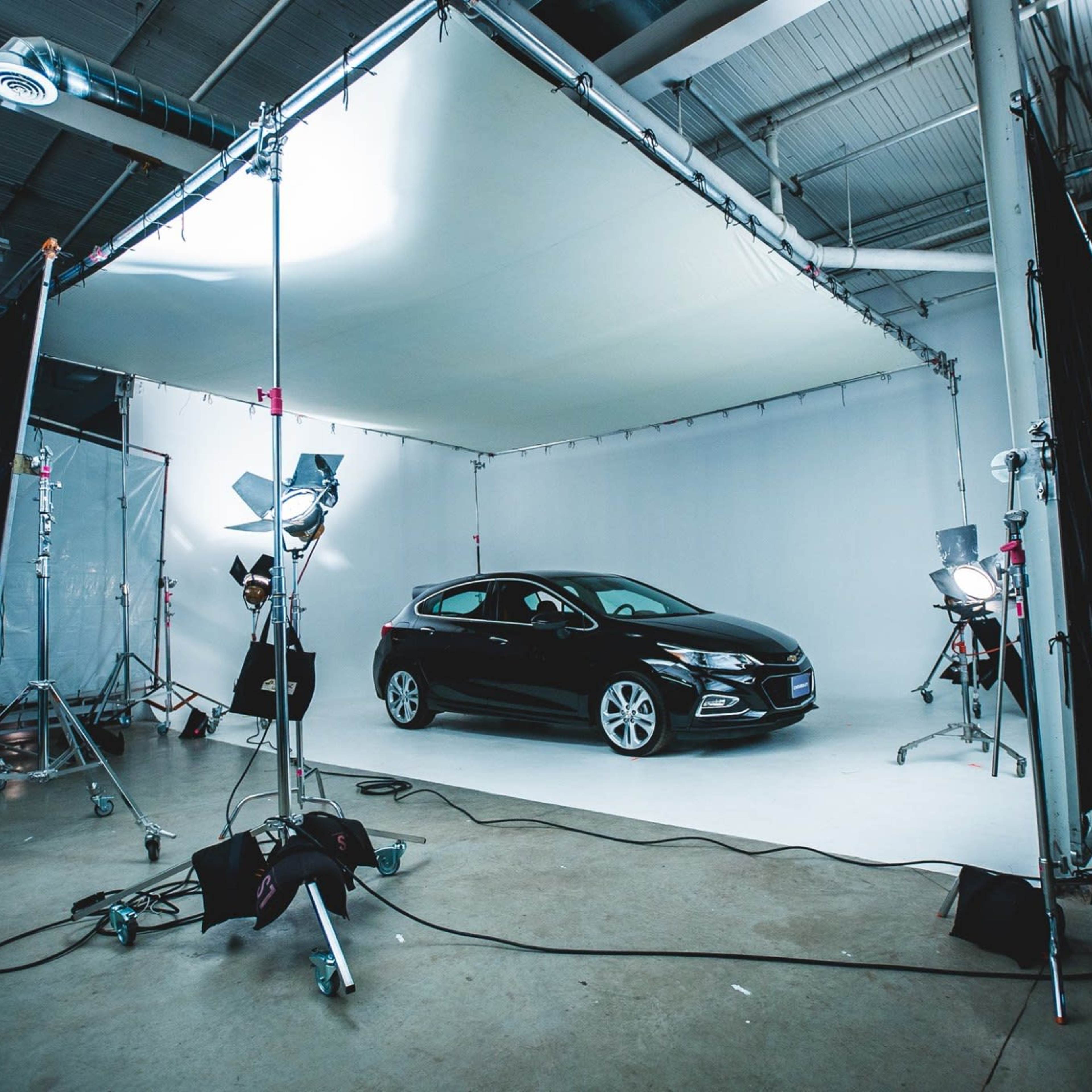 A black car is positioned in a well-lit studio on a seamless white backdrop, surrounded by photography equipment.