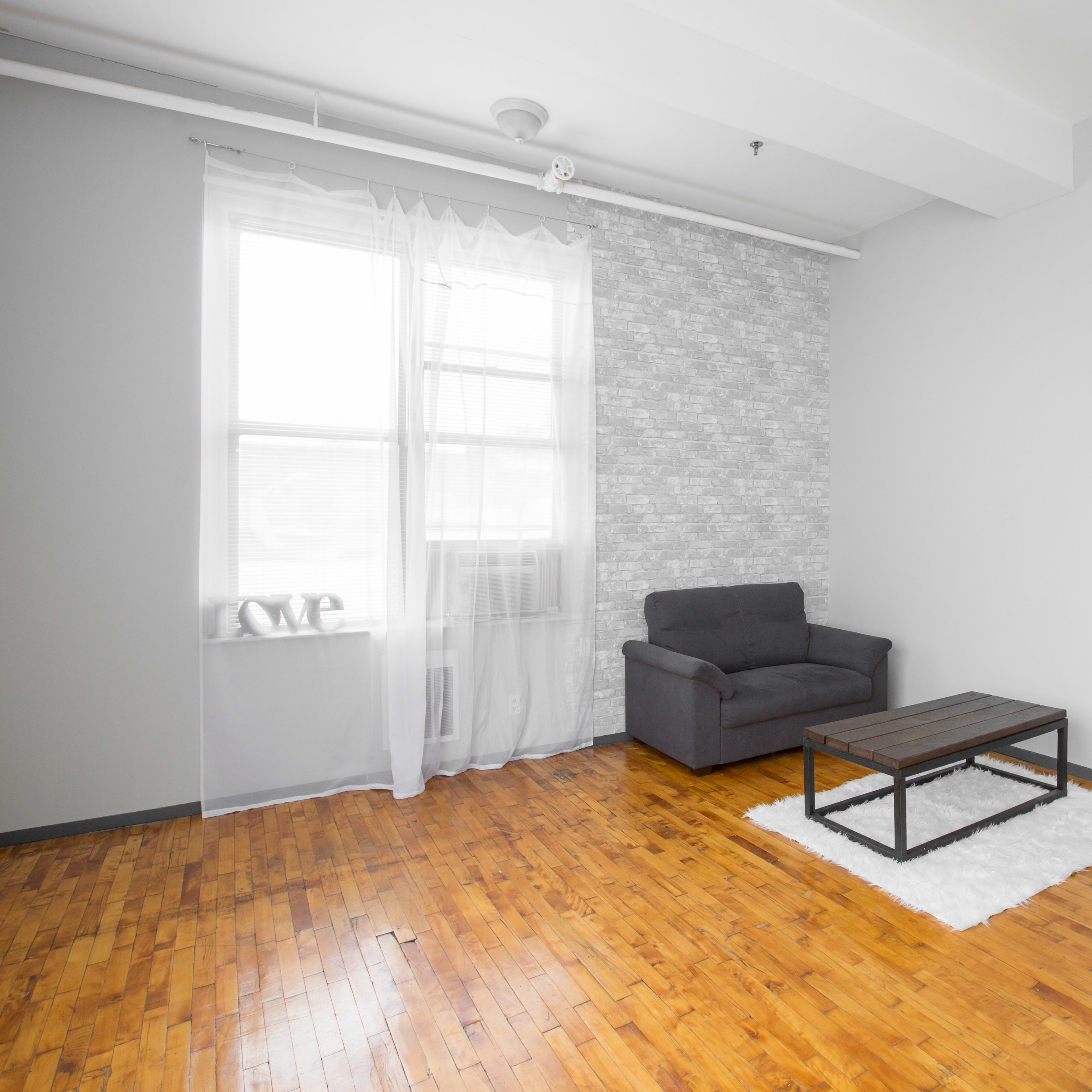 A minimalist living room features a gray chair, a wooden coffee table on a white rug, and a large window with sheer curtains.