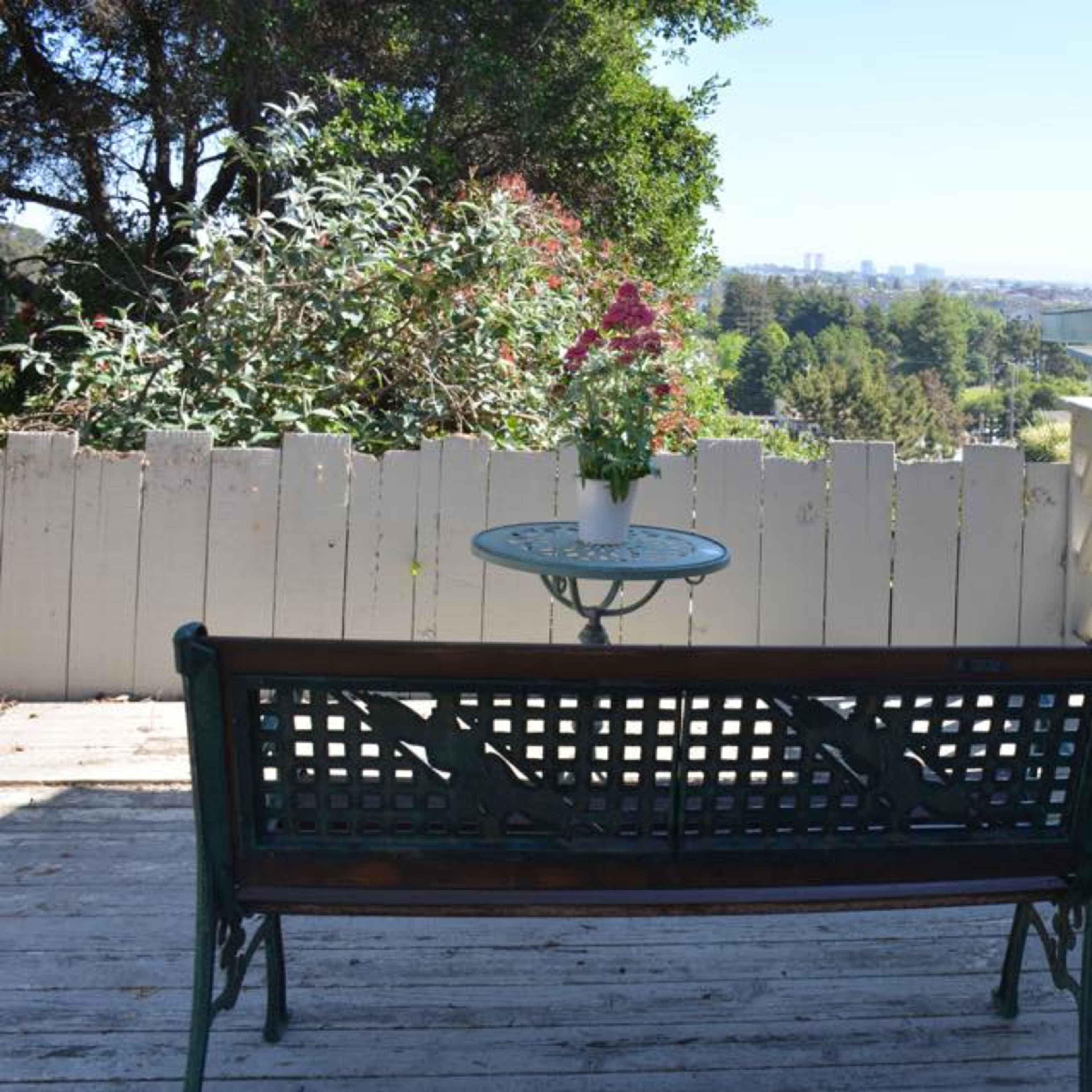 A green bench and a small table with a potted plant are positioned on a wooden deck overlooking a distant city skyline.