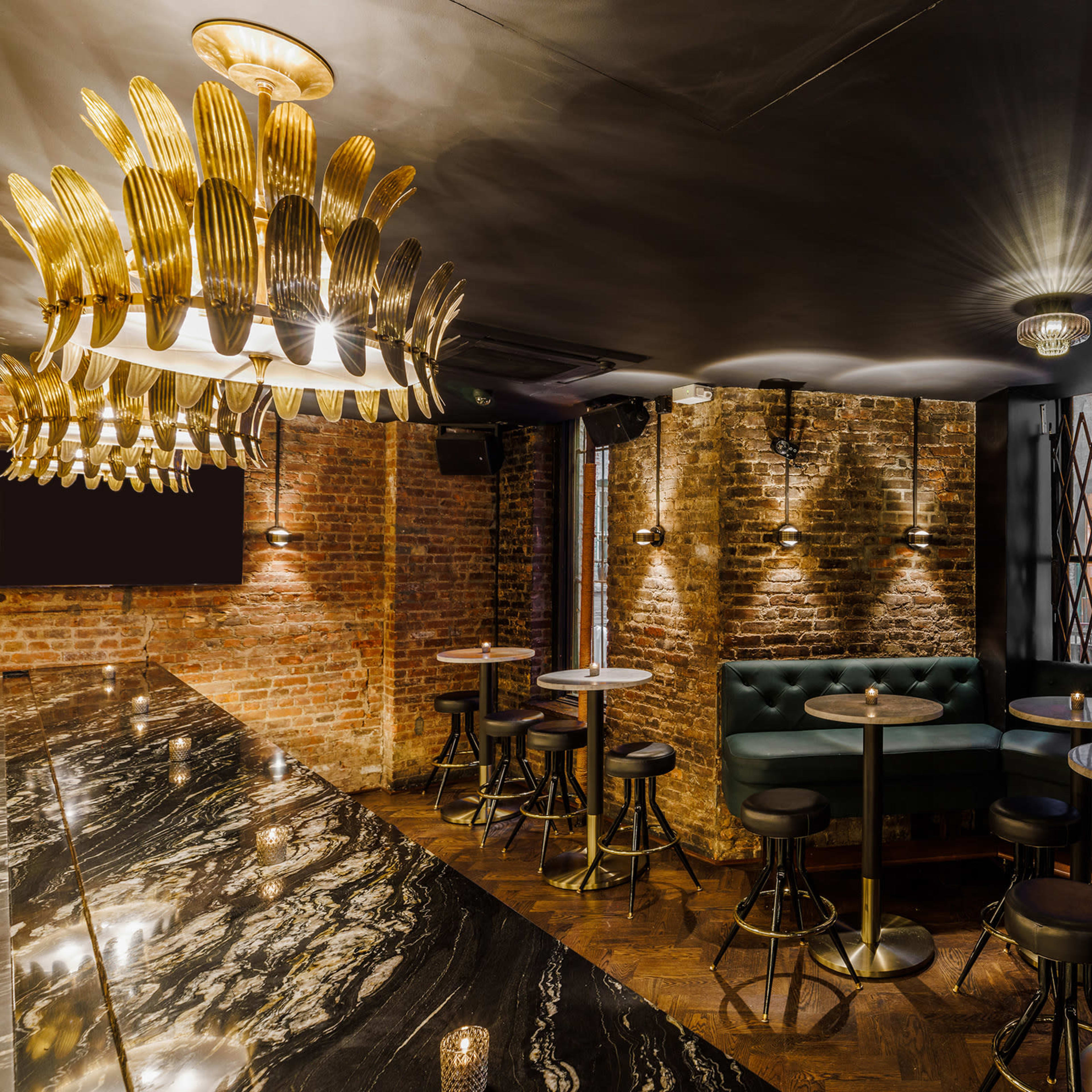 A stylish bar interior featuring brick walls, green seating, and a marble countertop with a decorative chandelier overhead.