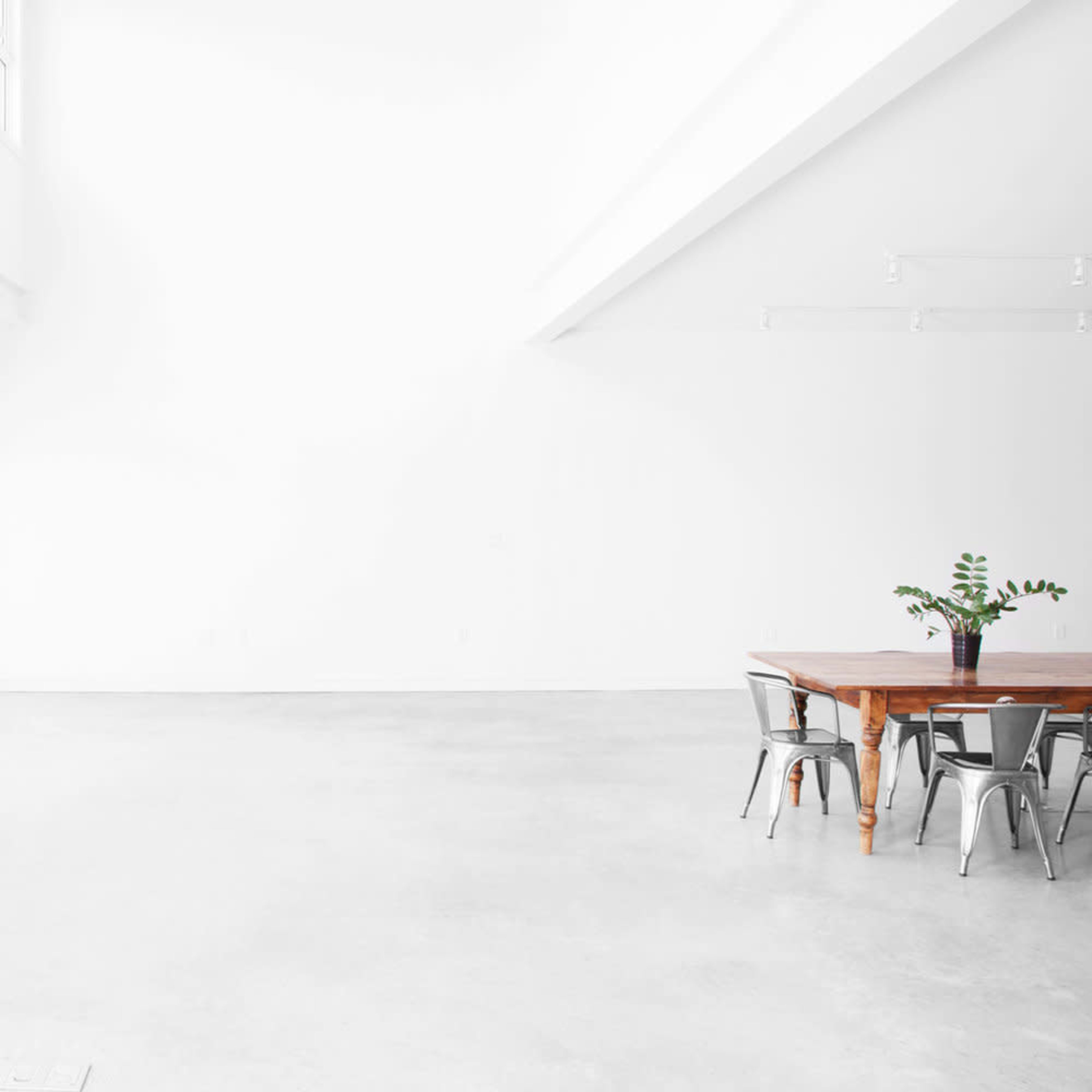 A spacious room with white walls and a polished concrete floor features a wooden table surrounded by metal chairs and large windows letting in natural light.