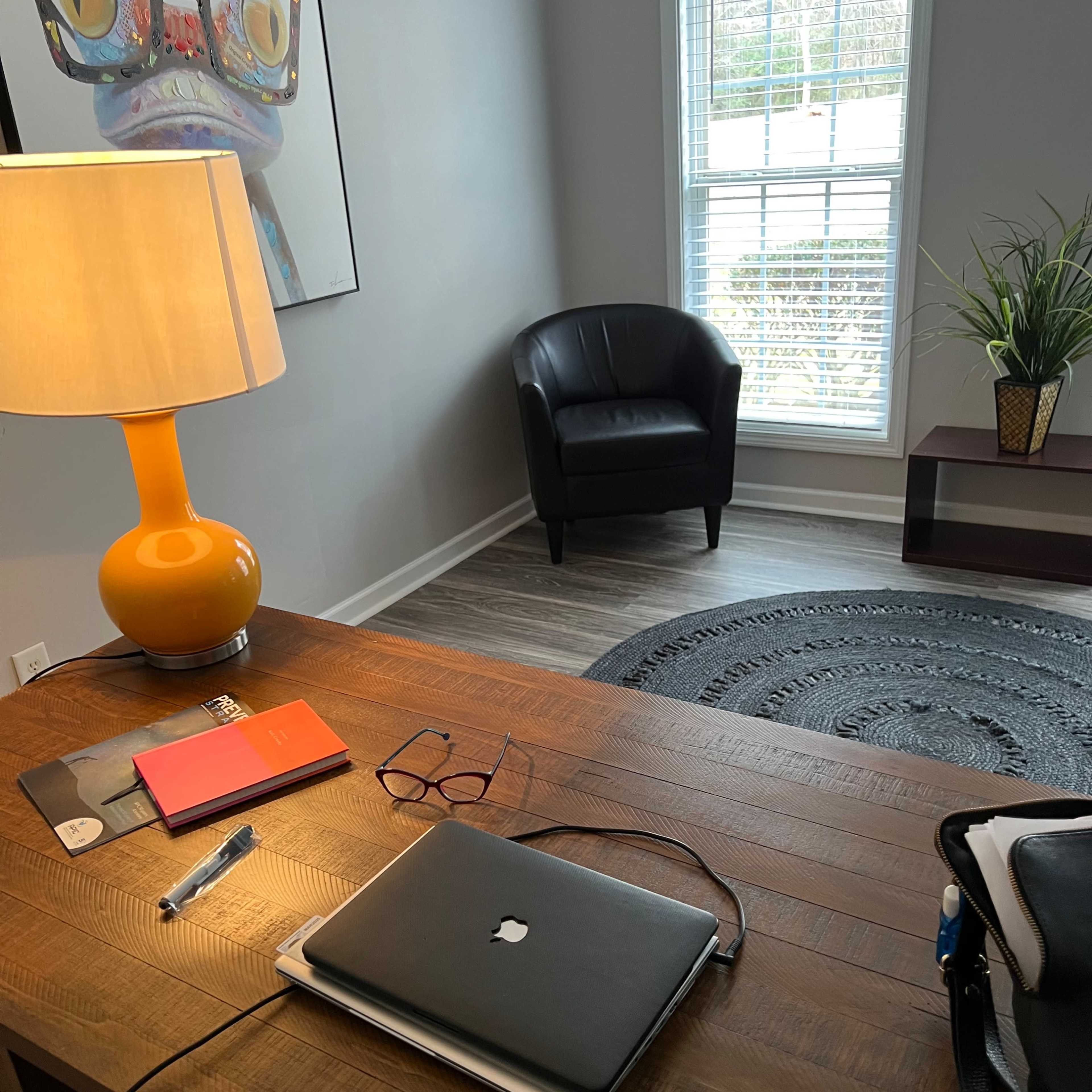 A wooden desk with a laptop, notebooks, and pens is positioned in a well-lit room featuring a chair, a lamp, and a wall art piece.