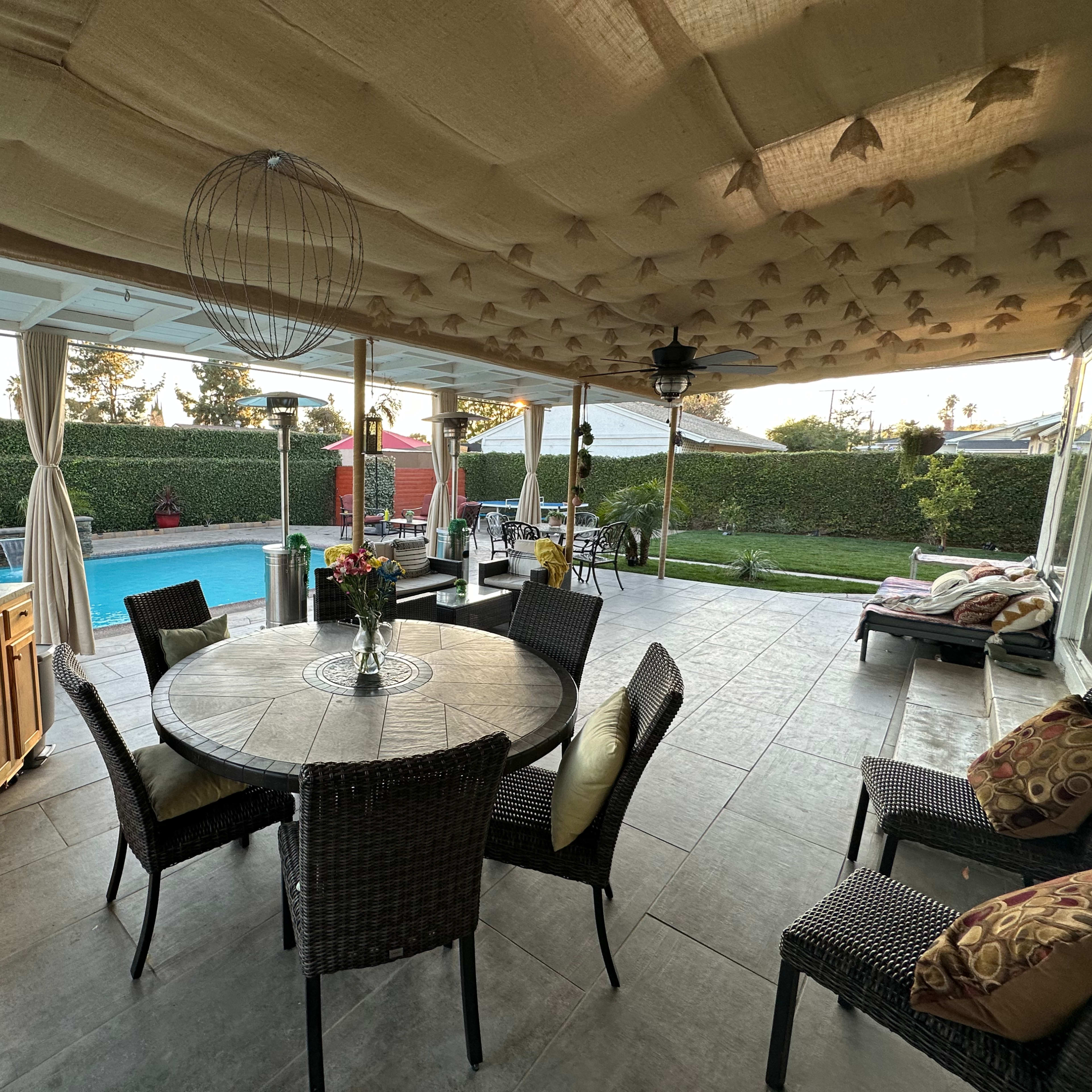 A covered patio area features a round dining table with wicker chairs, overlooking a backyard with a swimming pool and neatly trimmed hedges.